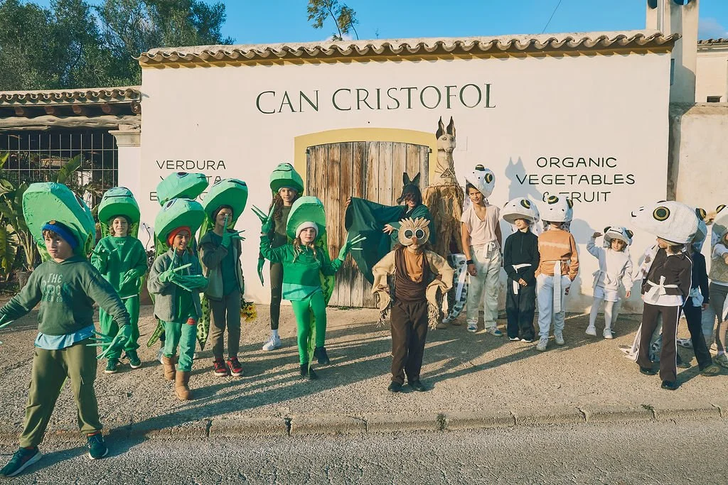 Niños disfrazados de plantas, vegetales y animales en una actividad escolar frente a un cartel que dice "Can Cristofol" y promueve verduras, frutas y vegetales orgánicos.