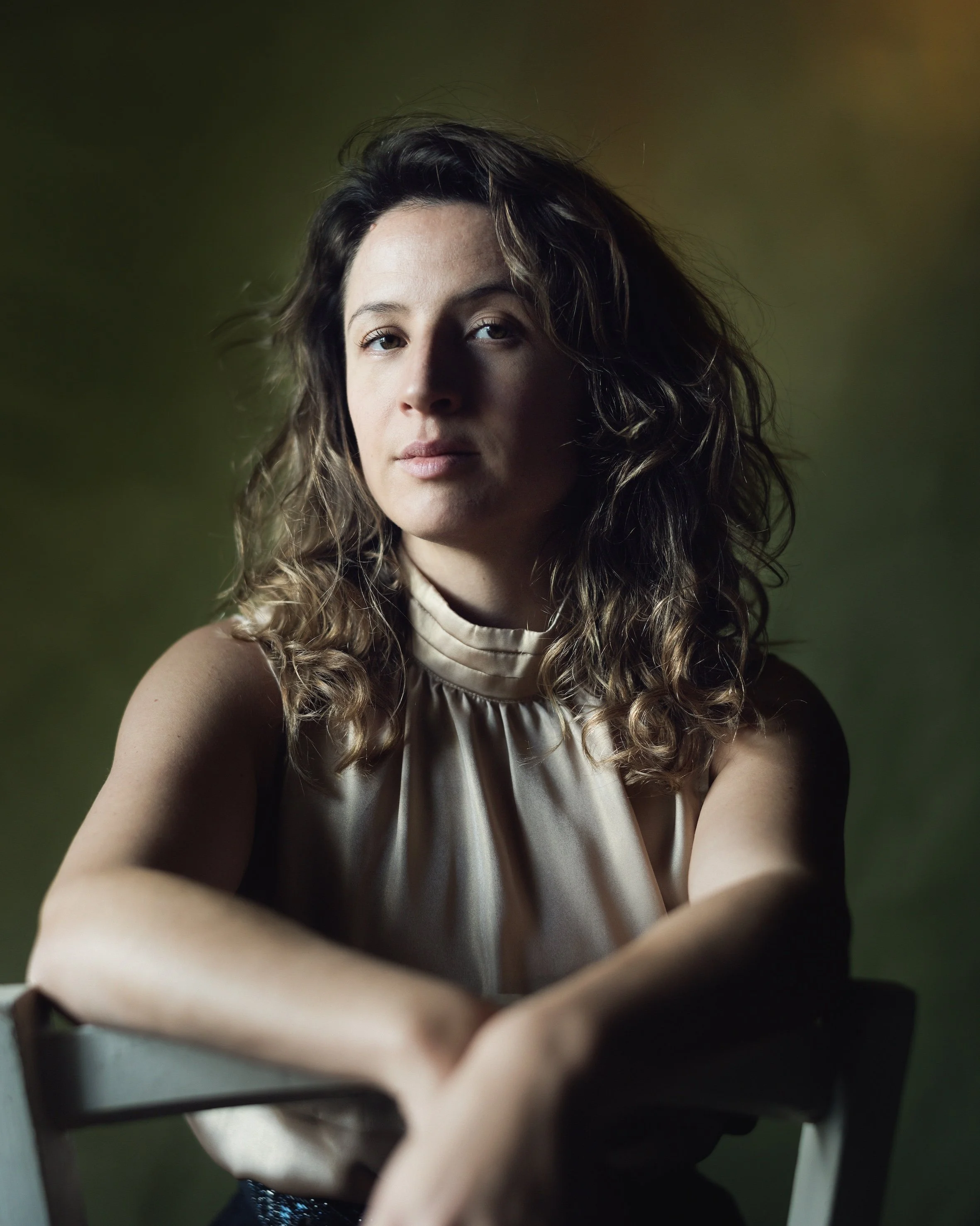 Retrato de una mujer con cabello rizado, usando blusa de satén color beige, con fondo de tonos oscuros y verdes difusos.