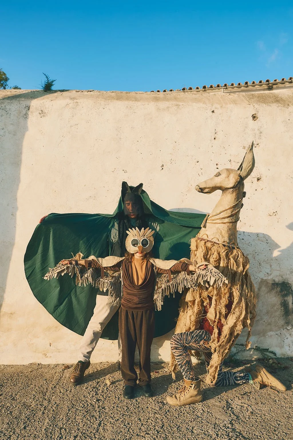 Tres personas en disfraces artísticos, una con disfraz de bosque y animal, otra con máscara de animal y otra con vestuario de león, en un fondo de pared blanca y cielo azul.