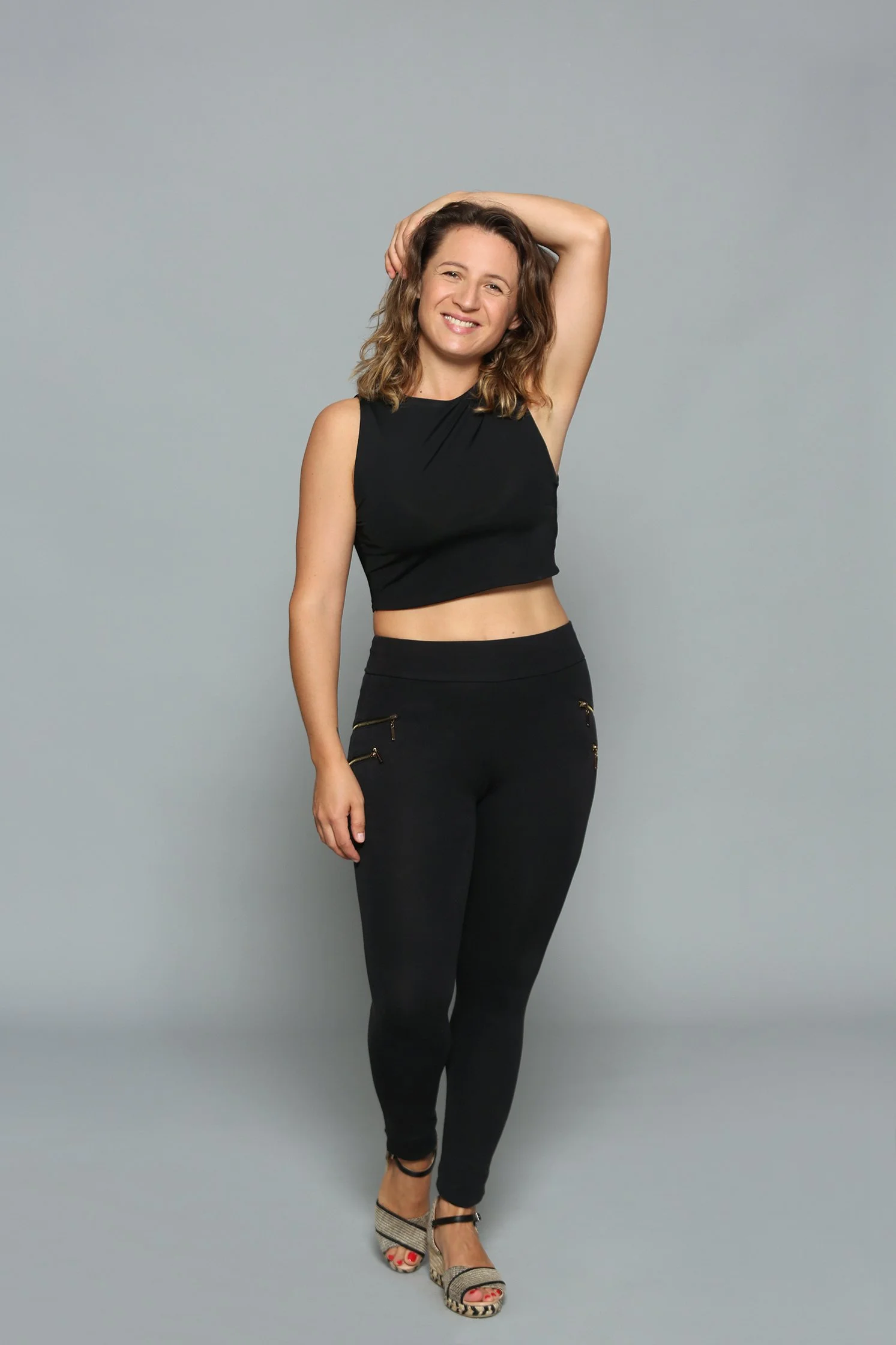 Mujer sonriente con cabello al hombro, vestido sin mangas negro y pantalones negros con cremalleras, posando con una mano detrás de la cabeza contra un fondo gris.