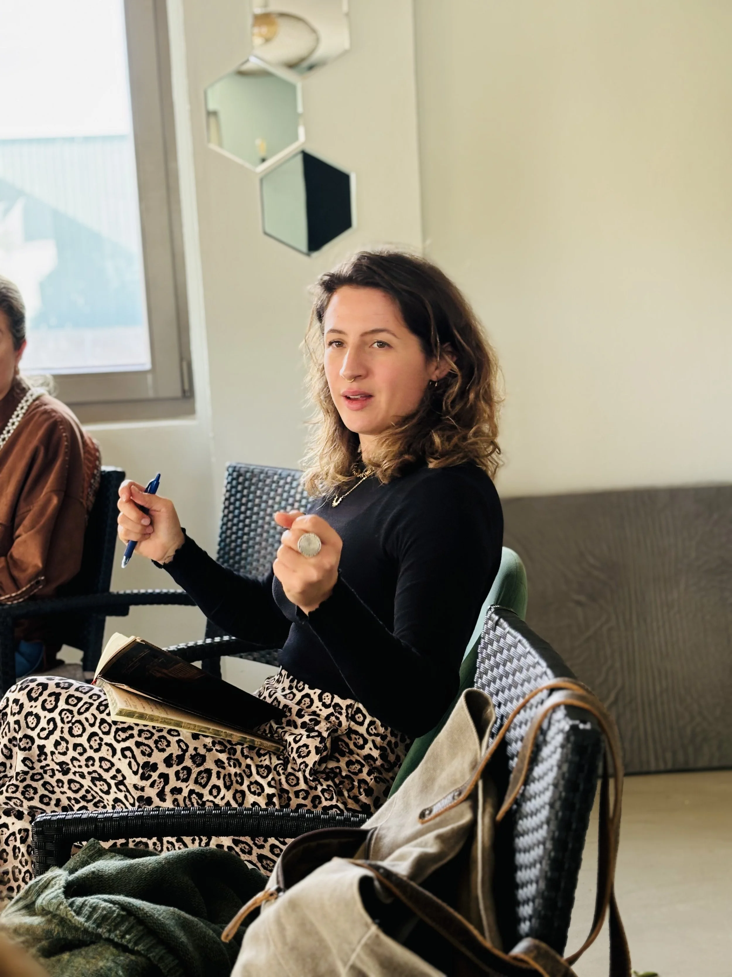 Mujer con cabello rizado y vestimenta de estampado de leopardo, sosteniendo un bolígrafo y un cuaderno en una reunión.