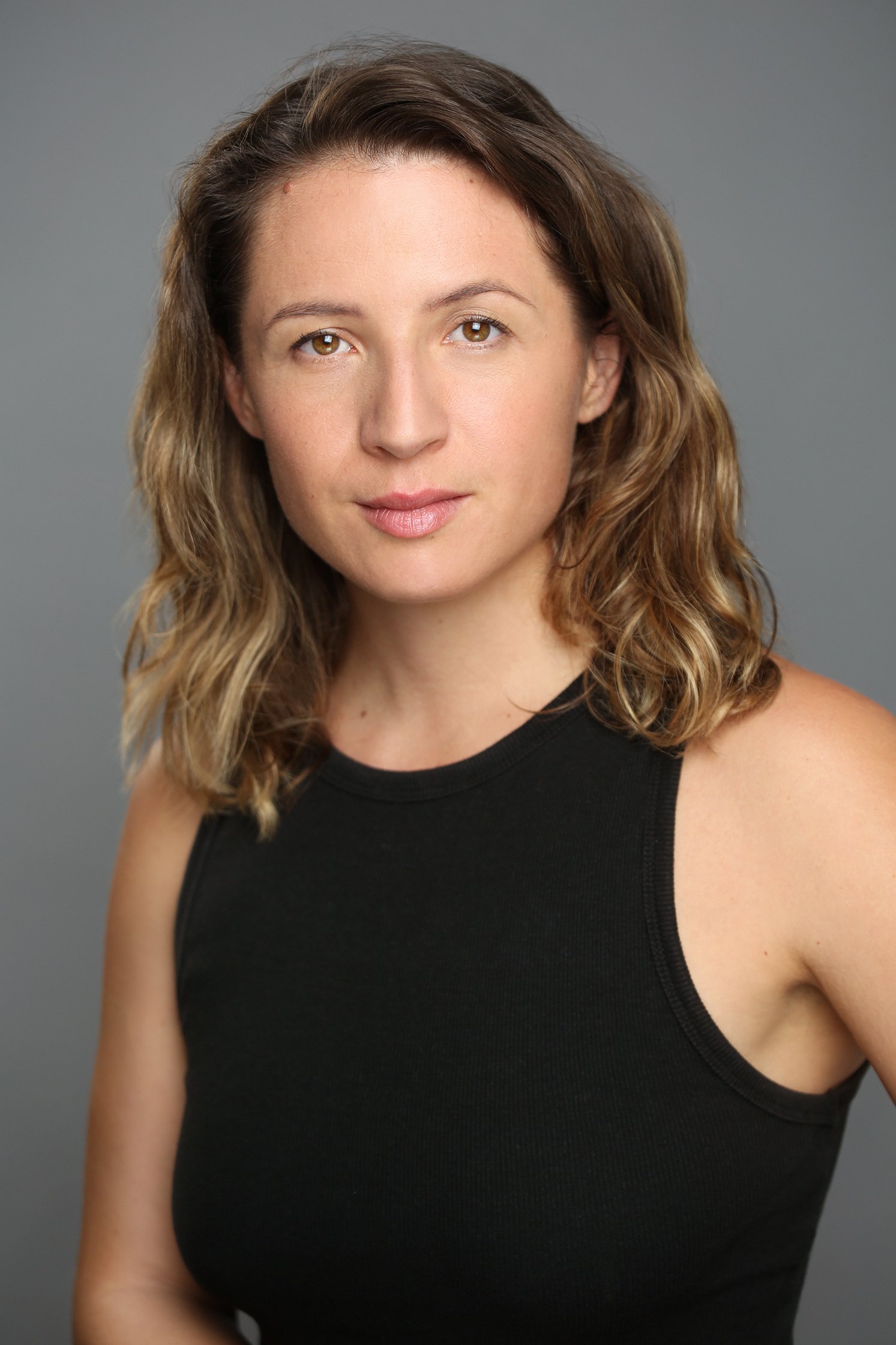 Retrato de una mujer con cabello castaño, cabello rizado y ojos claros, usando una camiseta sin mangas negra, contra un fondo gris.