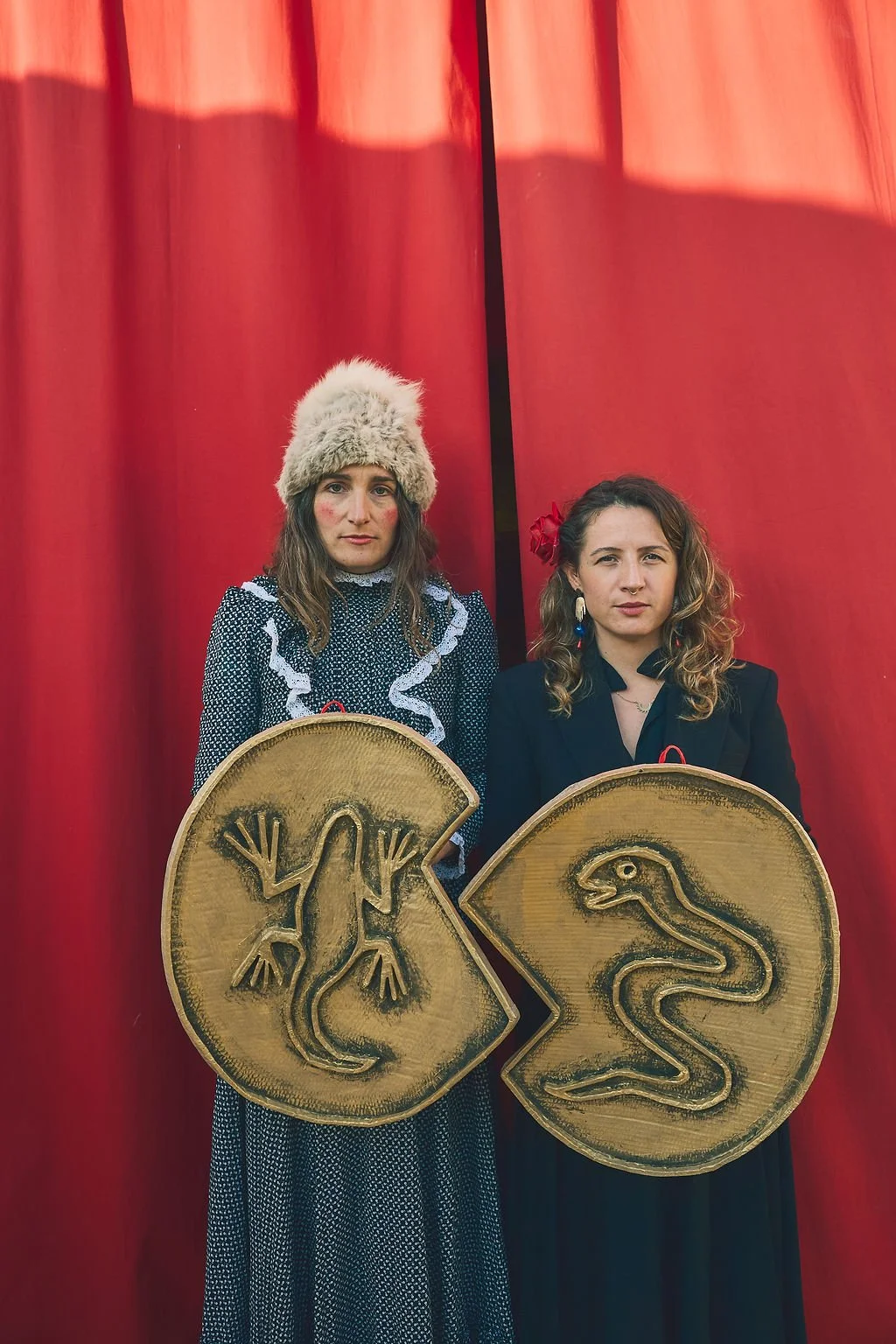 Dos mujeres sosteniendo escudos de madera con símbolos de animales, en un fondo de cortinas rojas.