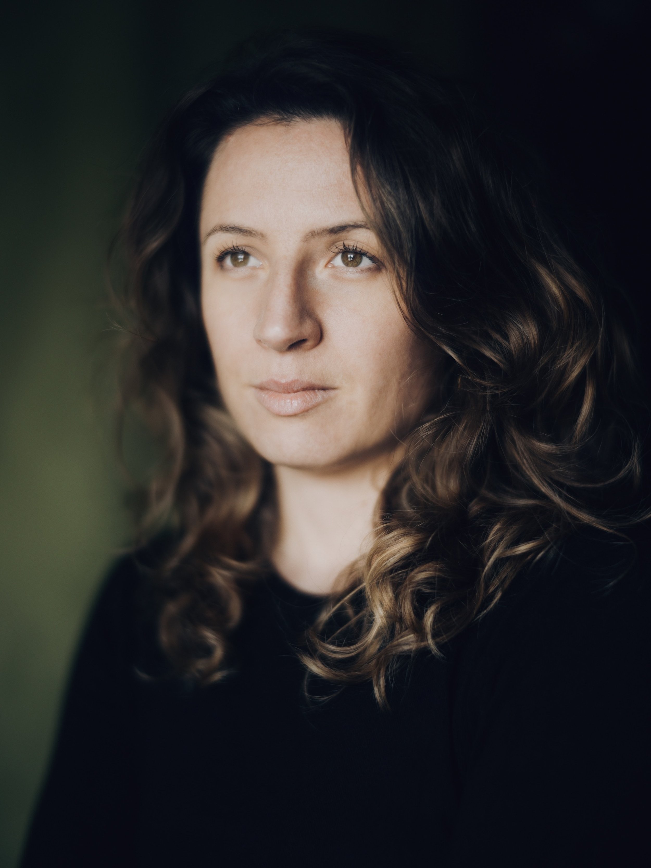 Retrato de una mujer con cabello rizado y oscuro, mirando hacia arriba y a la izquierda, con fondo oscuro y difuso.