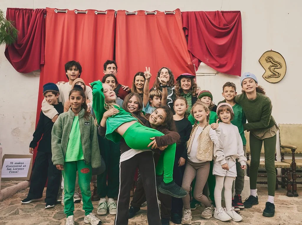 Grupo de niños y adultos posando en un escenario con cortinas rojas, algunos niños con ropa de colores vivos, en un ambiente que parece una obra de teatro o presentación escolar.