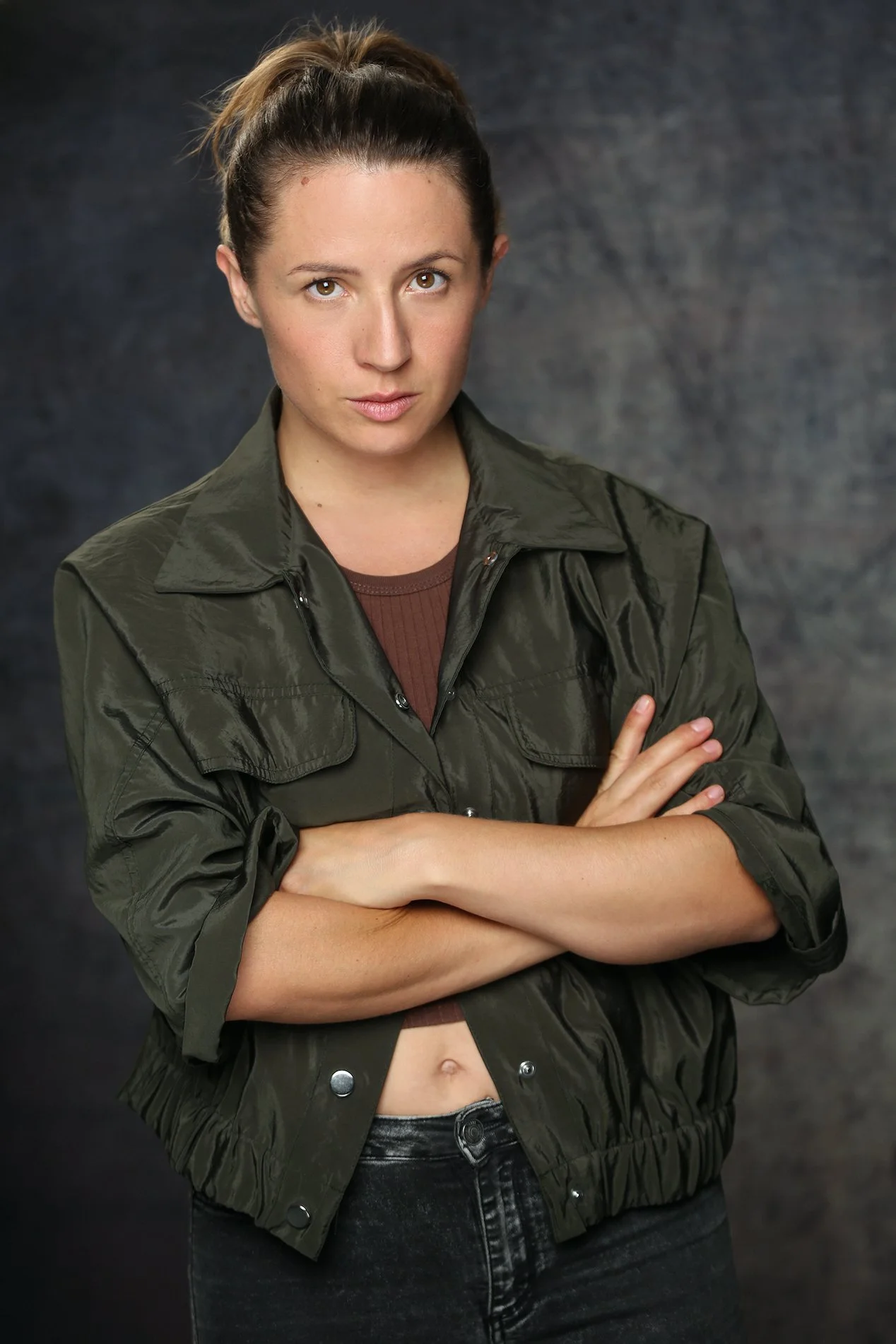 Joven mujer con cabello recogido, expresión desafiante, ojos mirando directamente, vestimenta casual con chaqueta verde oliva y pantalones oscuros, fondo oscuro y neutro.