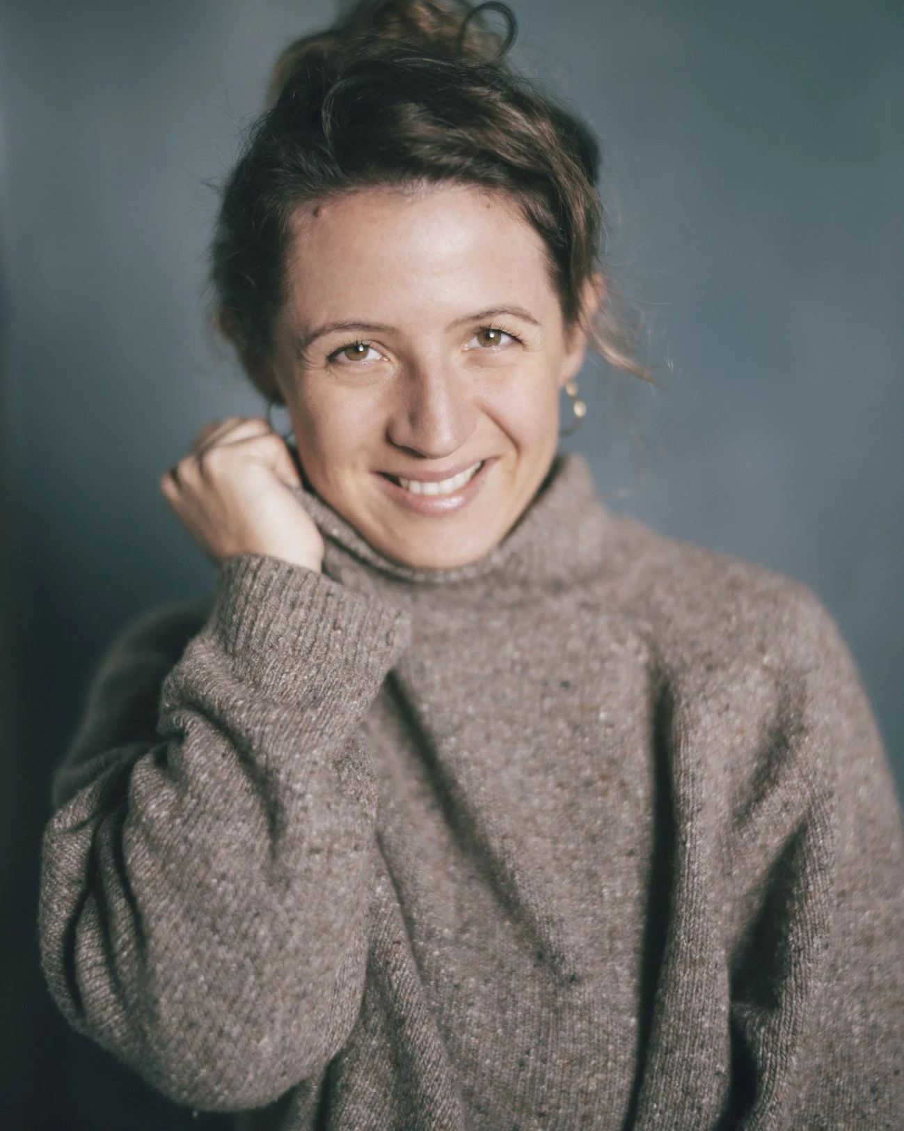 Retrato de una mujer joven con cabello castaño oscuro y rizado, sonriendo y usando un suéter de color marrón, en un fondo gris.