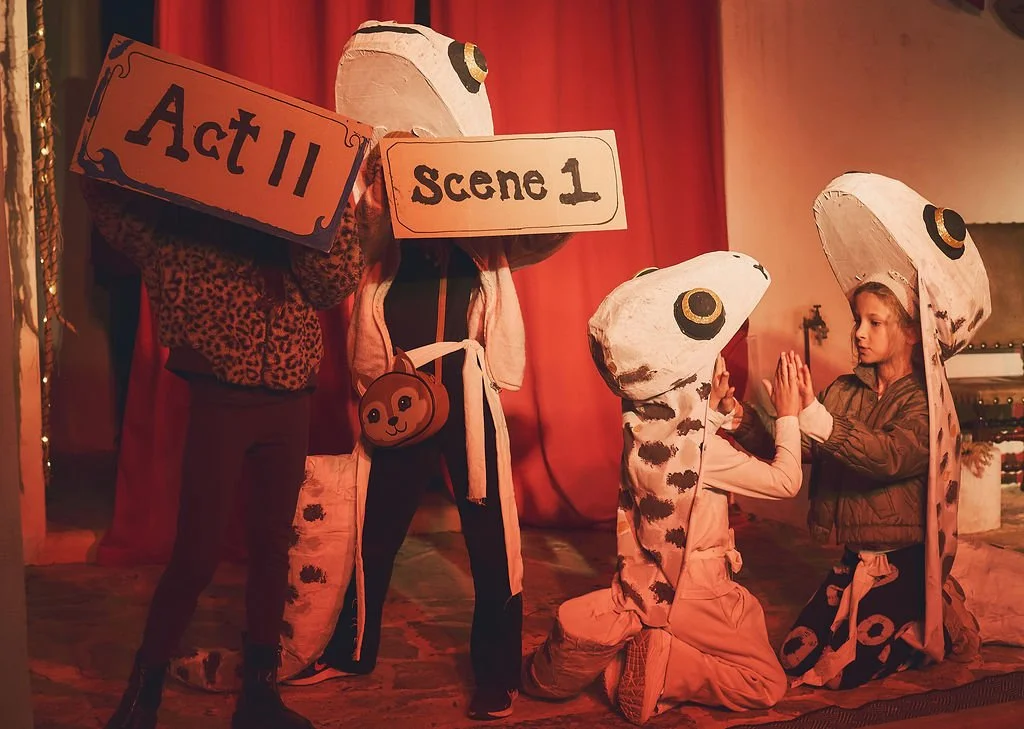 Dos niñas con disfraces de tortugas ninja en un escenario, simulando una escena teatral con letreros de 'Act II' y 'Scene 1', durante una actividad escolar o de teatro.