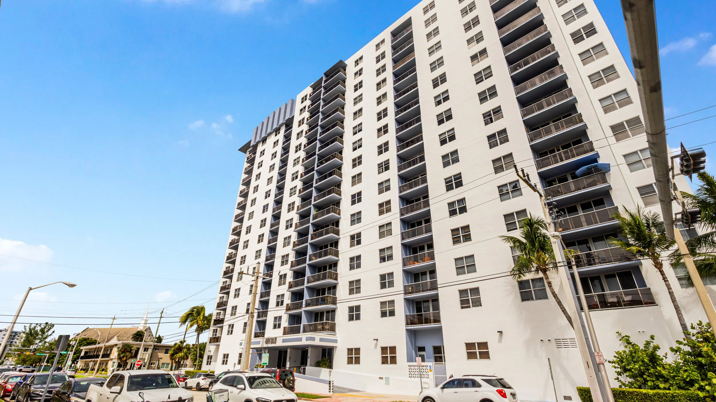 A tall white apartment building with multiple balconies set against a bright blue sky, with palm trees and parked cars in the foreground.