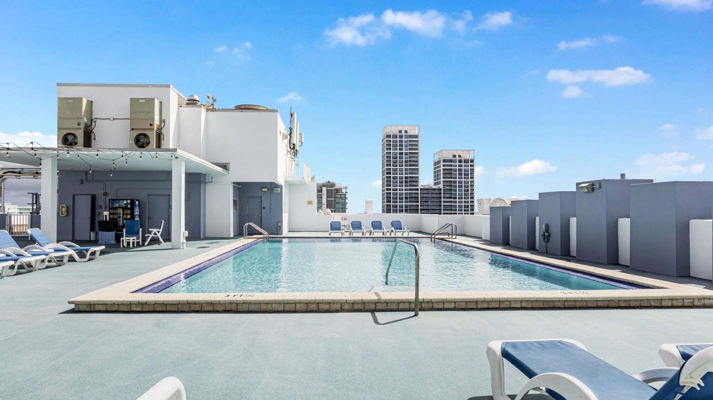 Rooftop swimming pool area with lounge chairs, a small shaded area, and city skyscrapers in the background under a blue sky with clouds.