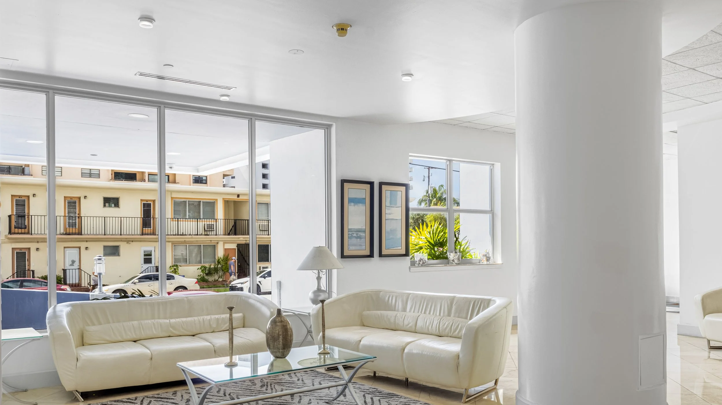 Bright lobby area with white leather sofas, a glass coffee table with decorative vases, a tall white lamp, and framed artwork on the wall. Large windows provide a view of neighboring apartment buildings and parked cars outside.