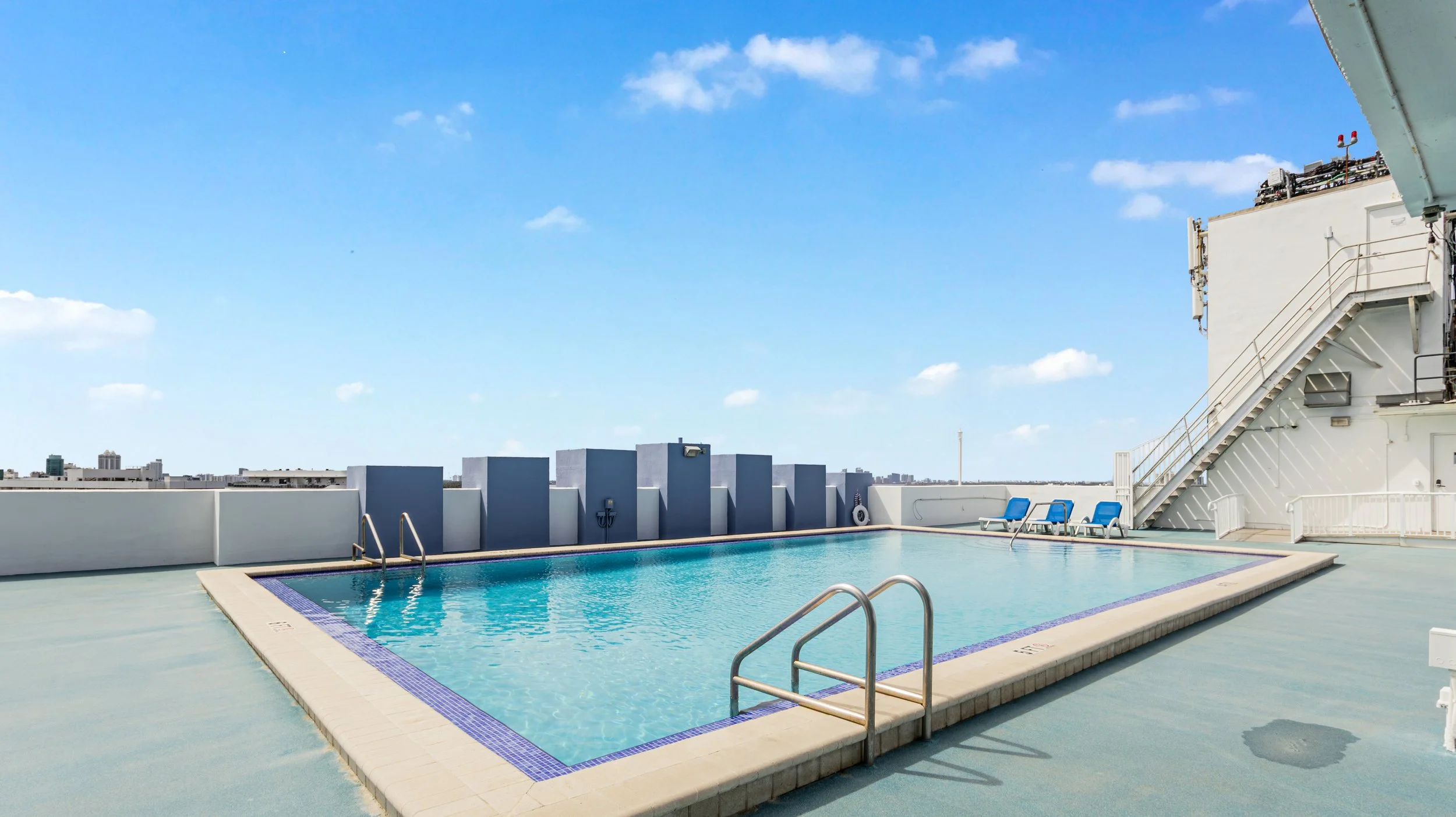 Rooftop swimming pool with blue lounge chairs and city skyline in the background under a partly cloudy sky.