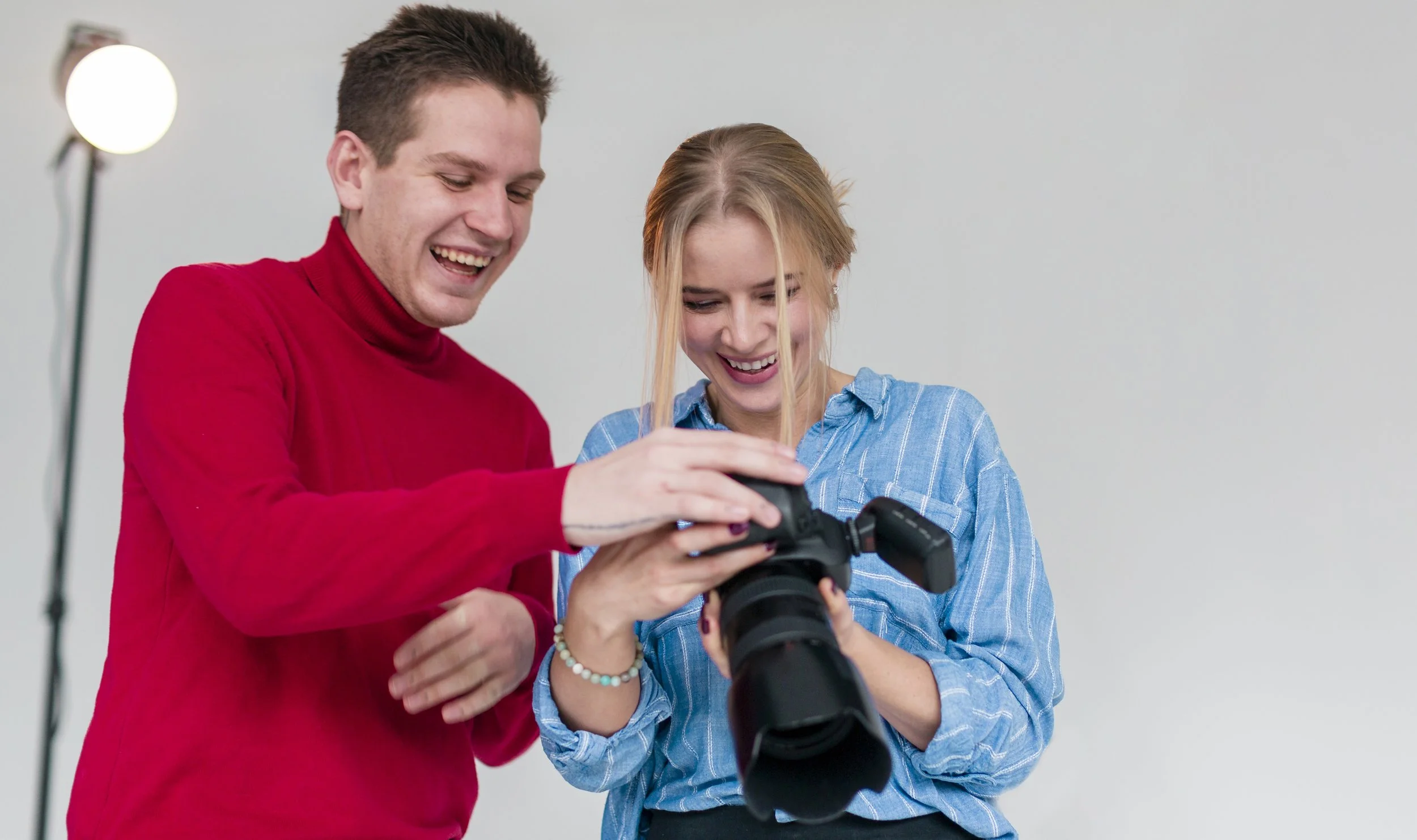 Two smiling people, a man in a red shirt and a woman in a blue shirt, are looking at the camera controls on a professional camera in a studio setting.