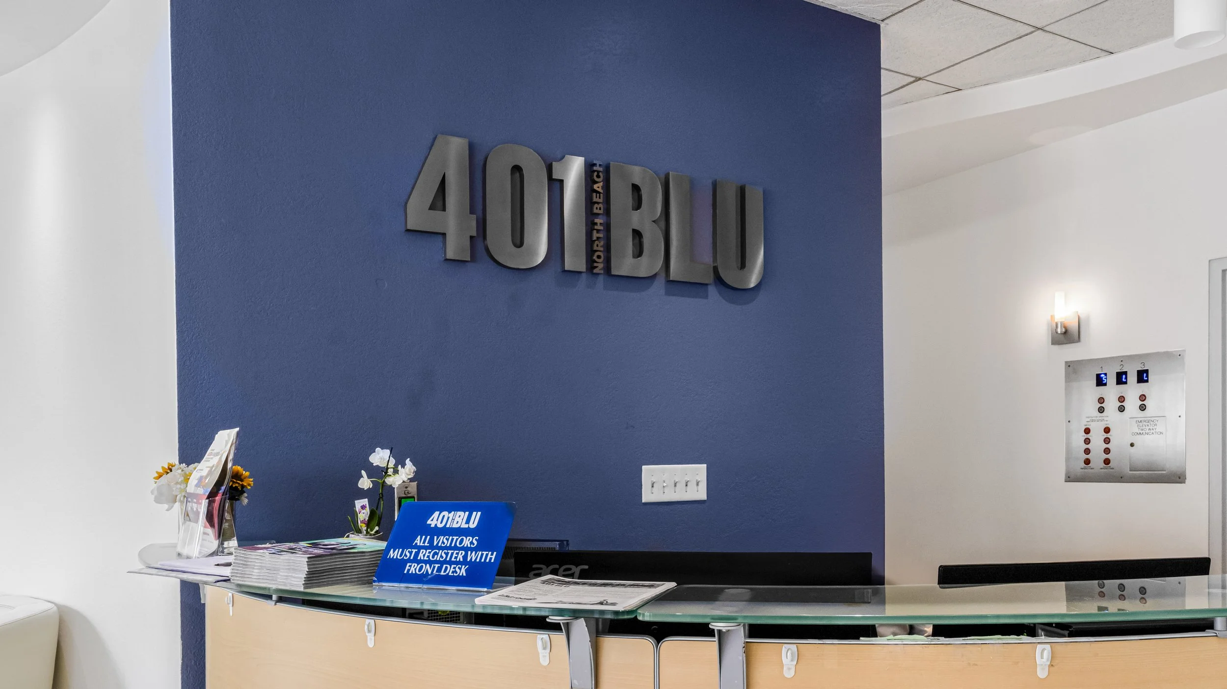 Reception area with a blue and white wall, a sign reading '401 BLU NORTH BEACH,' a desk with brochures, a blue sign stating 'ALL VISITORS MUST REGISTER WITH FRONT DESK,' and a communication panel on the wall.