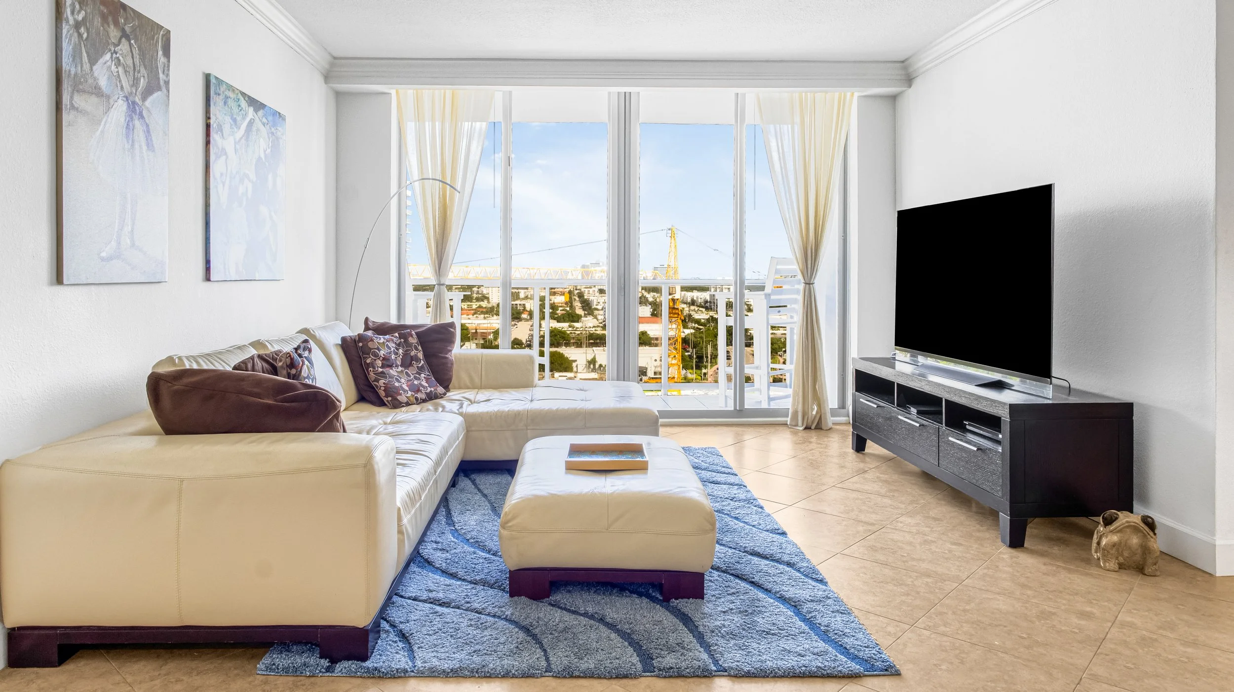 Living room with a cream sectional sofa, decorative pillows, a matching ottoman, a blue rug, a large flat-screen TV on a black TV stand, a sliding glass door with light curtains, a city view outside, and wall art.