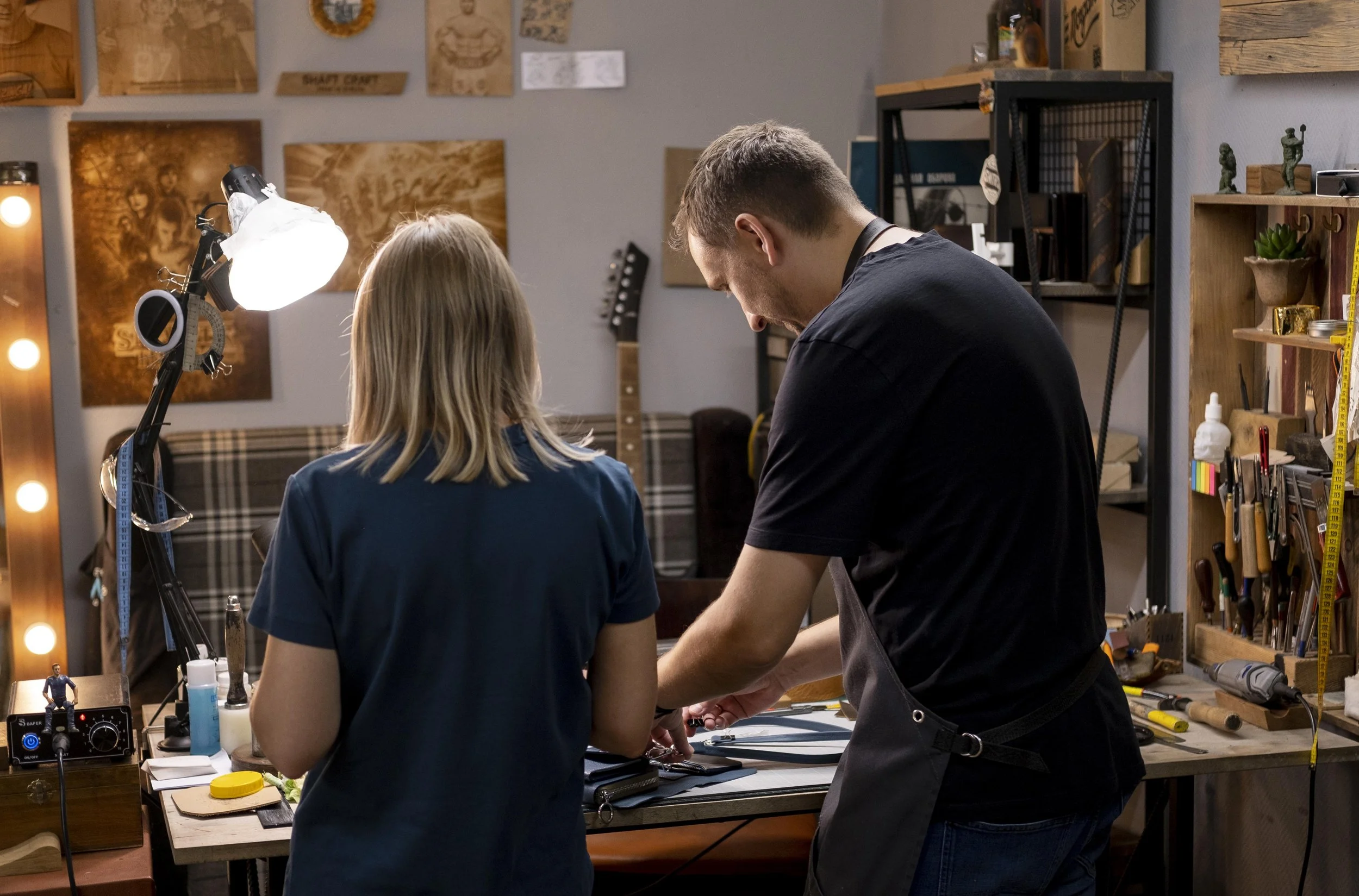 Two people working on a craft project in a workshop with tools and artwork on the walls.
