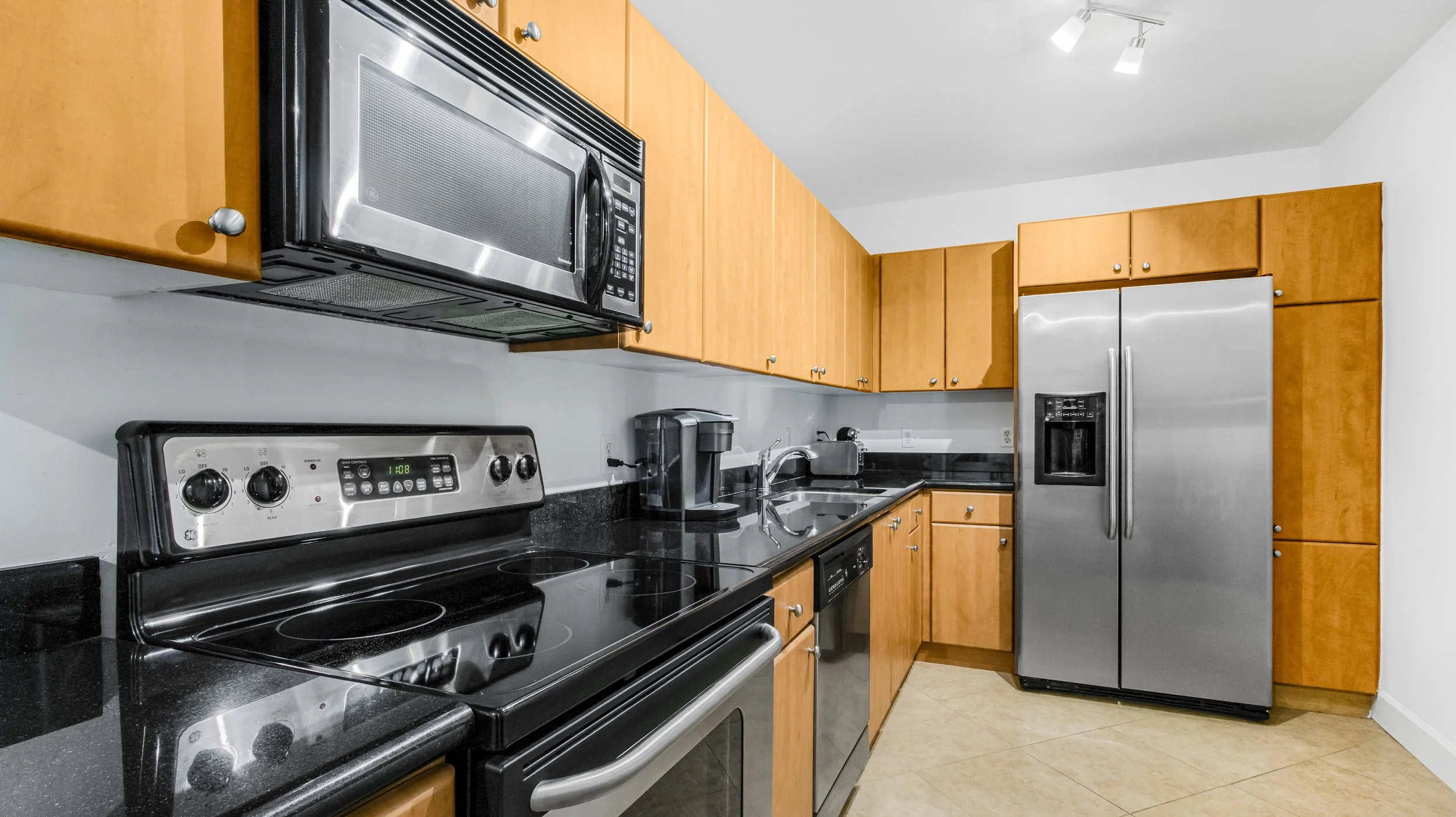 Kitchen with black appliances, including a microwave above a stove, a coffee maker, a sink, and a refrigerator. Wooden cabinets with silver knobs are mounted on the walls, and the floor is tiled.