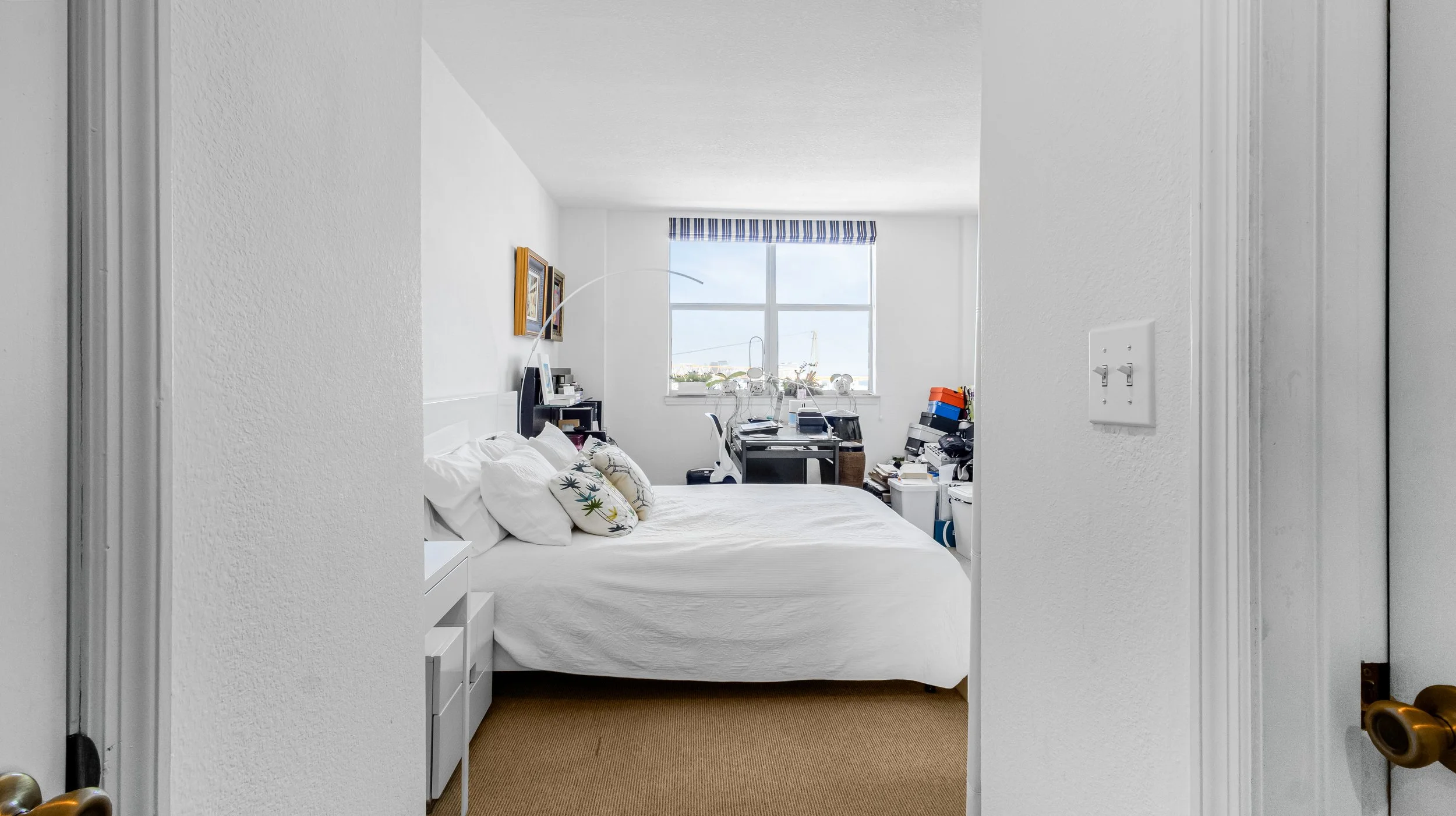 View of a bedroom with a bed, pillows, a nightstand, a window with a striped valance, and a cluttered desk and shelves in the background.