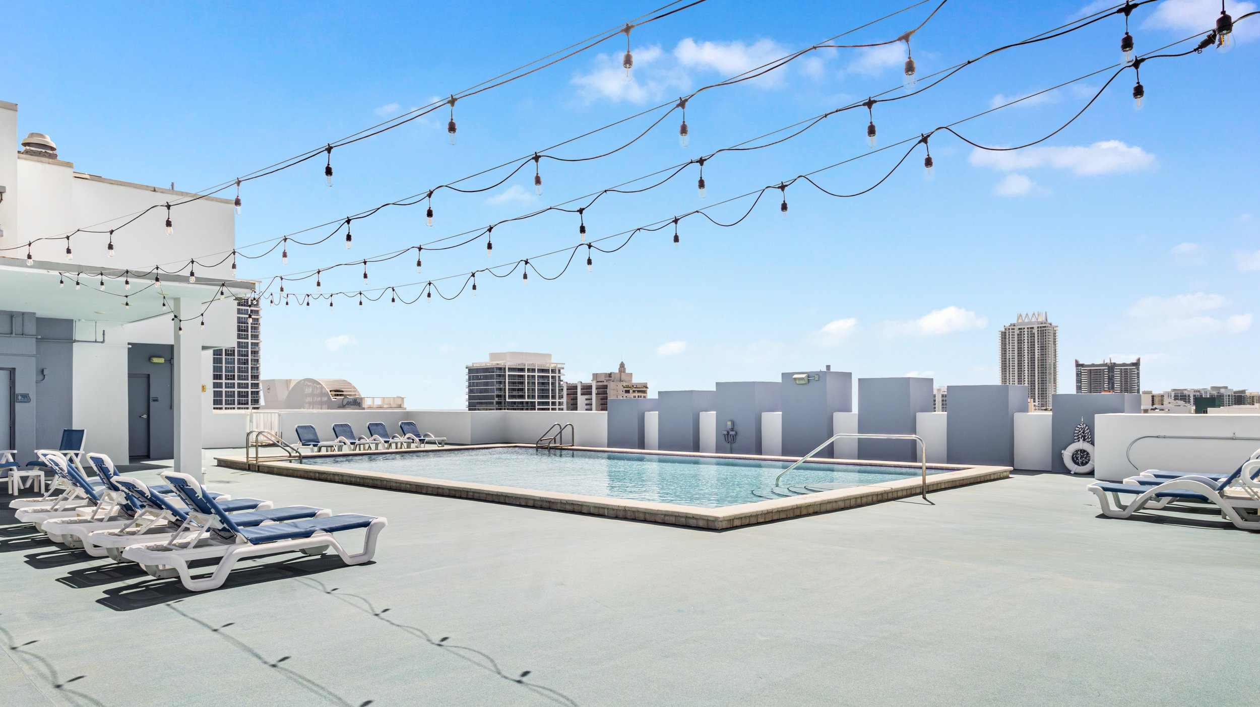 Rooftop swimming pool under a blue sky with city buildings in the background, string lights overhead, and several lounge chairs around the pool.