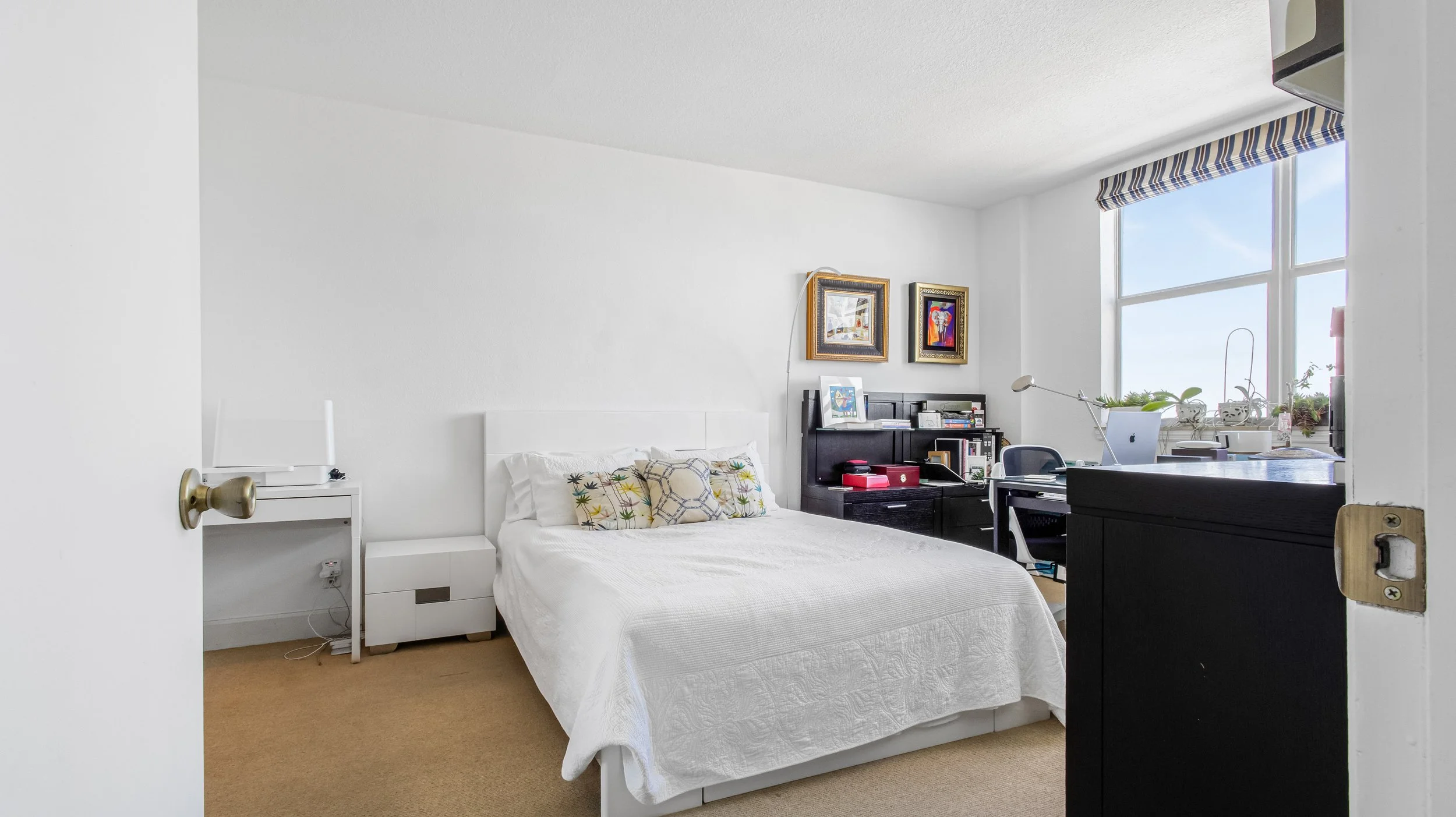 A clean, minimal bedroom with a white bed, decorative pillows, a black bookshelf with art and books, a desk with a computer, and a large window with a view outside.