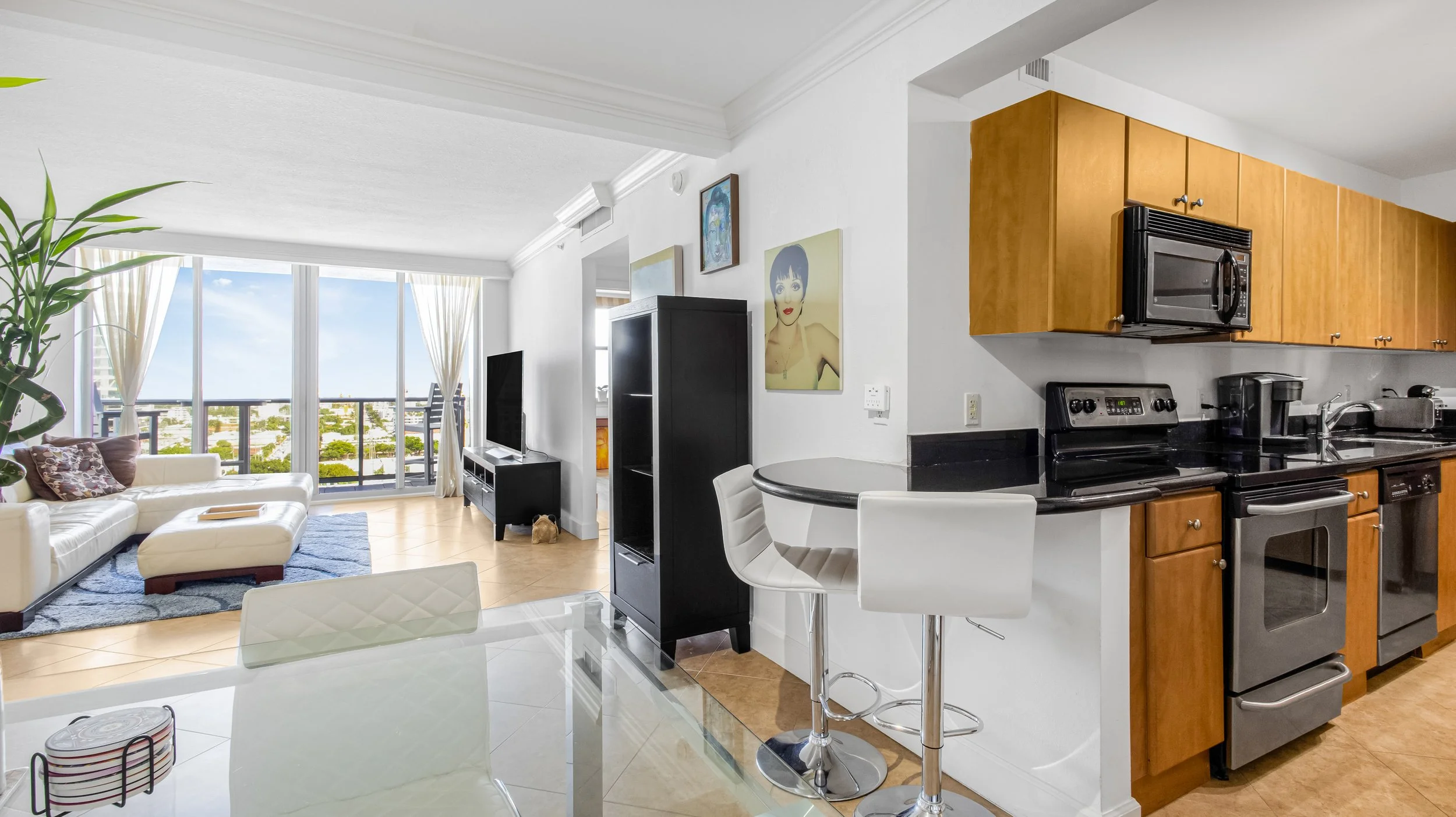 Modern open-concept living room and kitchen with large windows, white couch, black TV stand, black entertainment center, artwork, and kitchen area with wooden cabinets and black appliances.