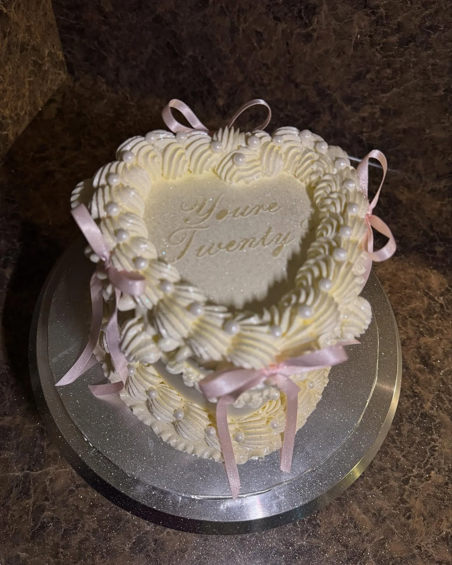 A two-tiered, cream-colored birthday cake with pink ribbons and pearl decorations, inscribed with the words 'Your Twenty' on the top tier.
