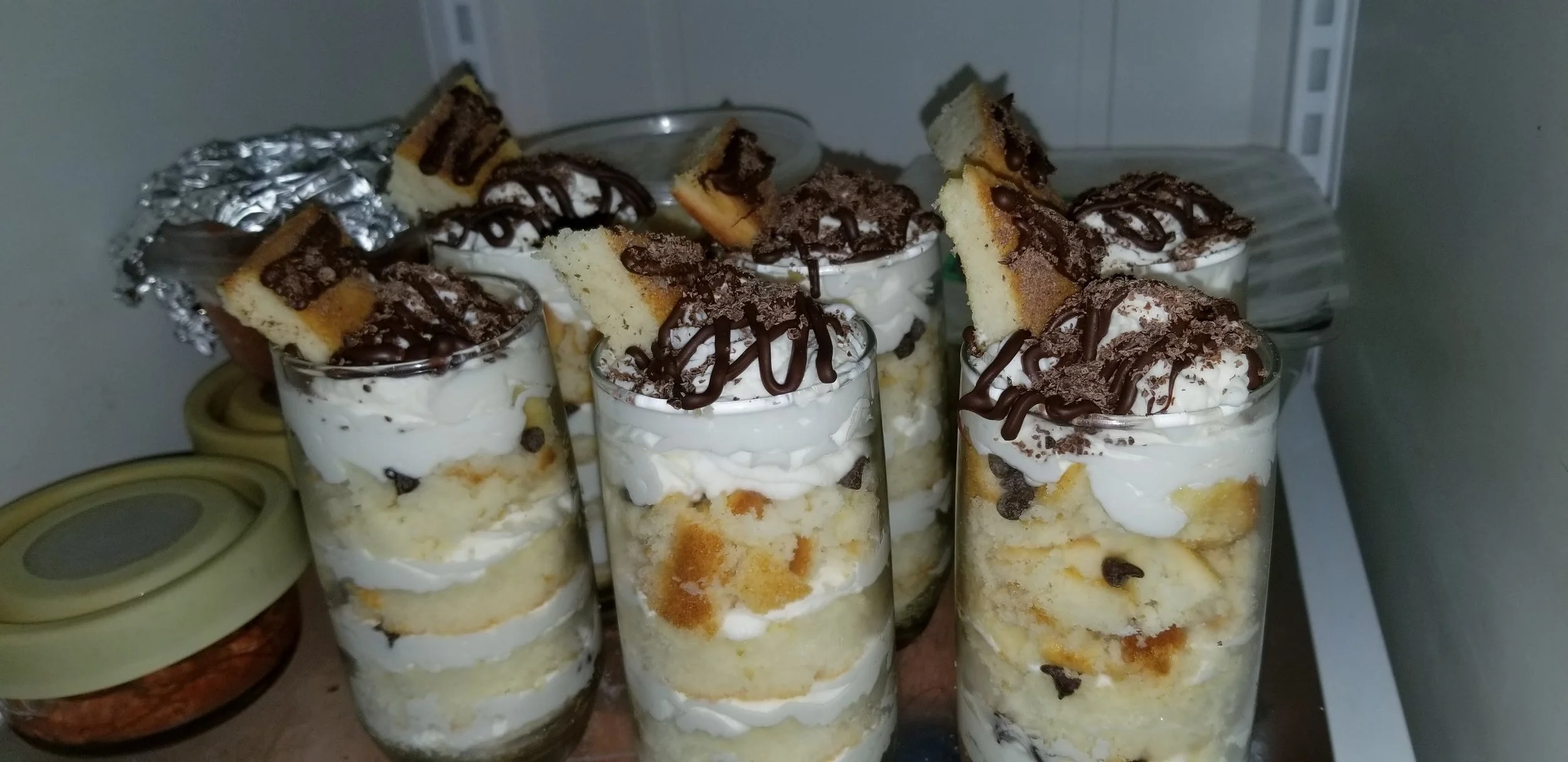 Several dessert parfaits in tall glasses with layers of cake, whipped cream, chocolate chips, and topped with graham crackers, chocolate drizzle, and crumbled cookies.