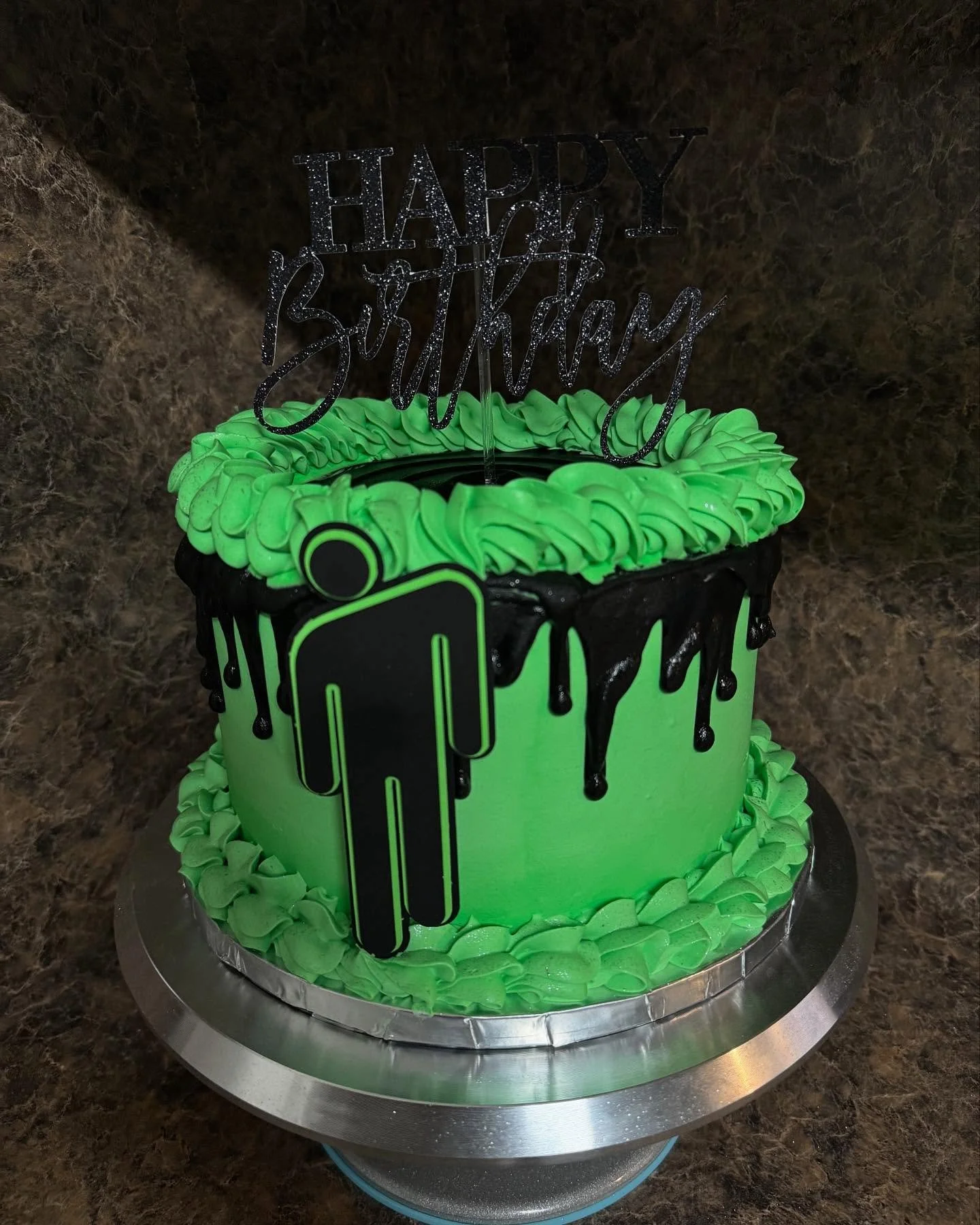 A birthday cake with green icing and black drips, topped with a glittery 'Happy Birthday' sign, and featuring a black silhouette of a person on the side.