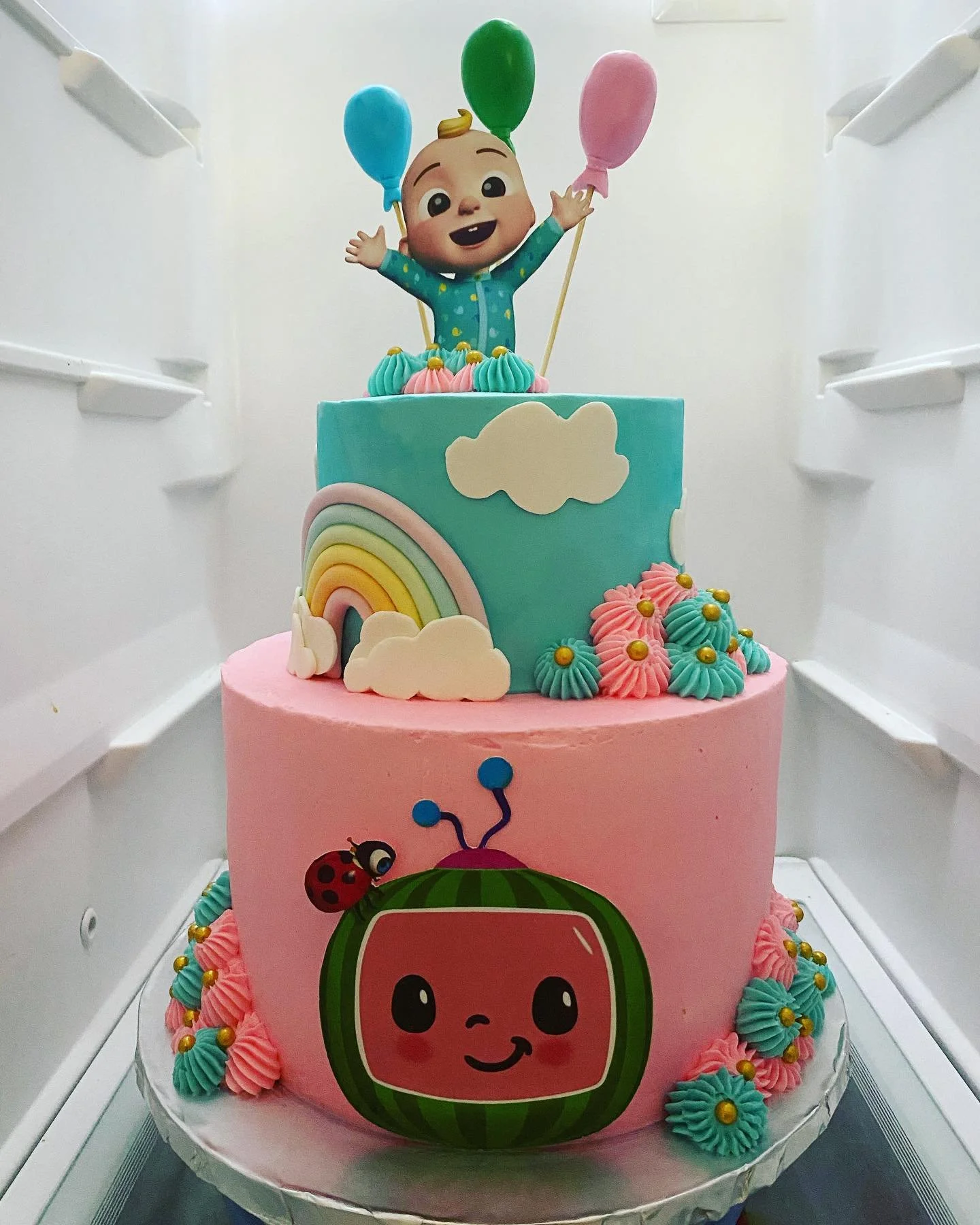 Colorful two-tier birthday cake with cartoon characters, rainbows, clouds, and balloons decorations, inside a fridge.