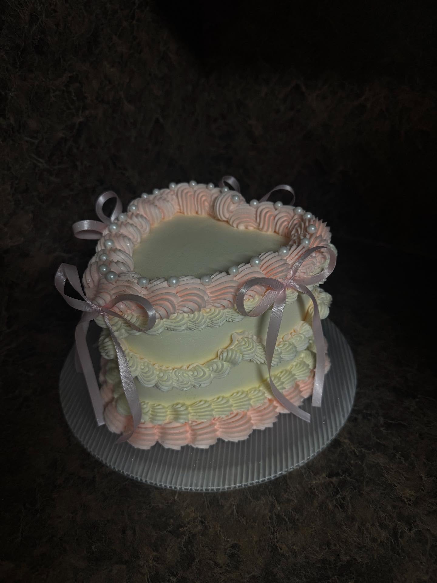 A two-tiered cake with pastel pink and cream-colored frosting decorated with pearl-like beads and satin ribbons.