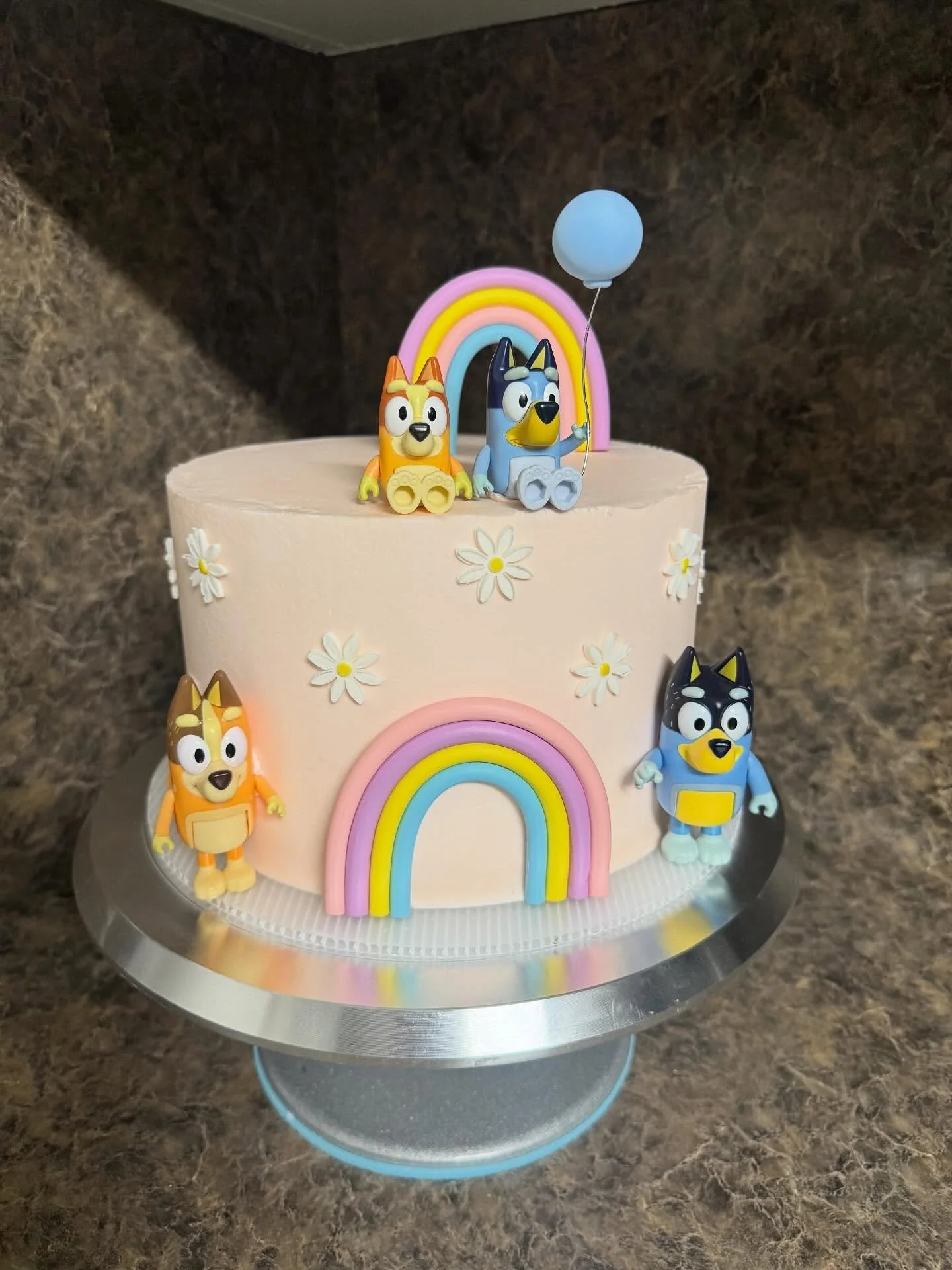 A pink birthday cake decorated with colorful rainbows, white daisies, and figurines of characters from the children's show Bluey. The cake has three figurines and an additional one on the side, along with a blue balloon. The cake is placed on a silve