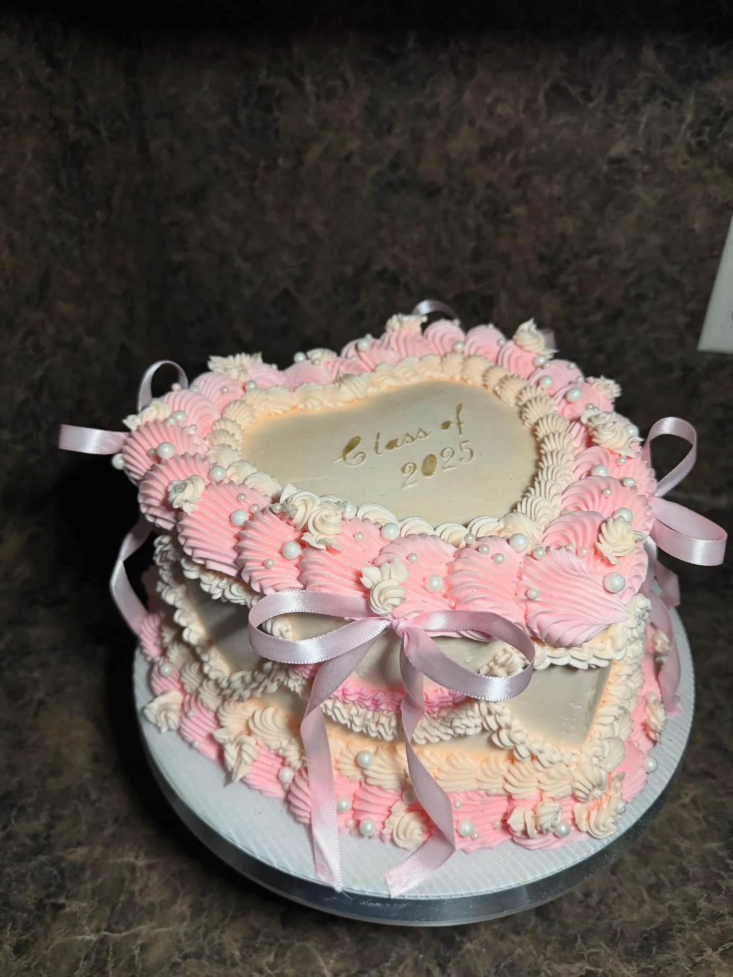 Two-tiered graduation cake decorated with pink and white icing, pearl-like sugar beads, and pink ribbons, with the writing "Class of 2025" on the top layer