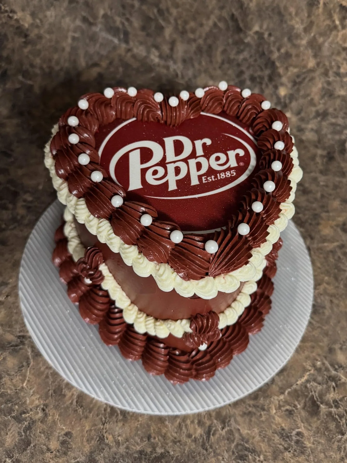 A heart-shaped cake with the Dr Pepper logo on top, decorated with white and chocolate frosting and small white edible pearls.