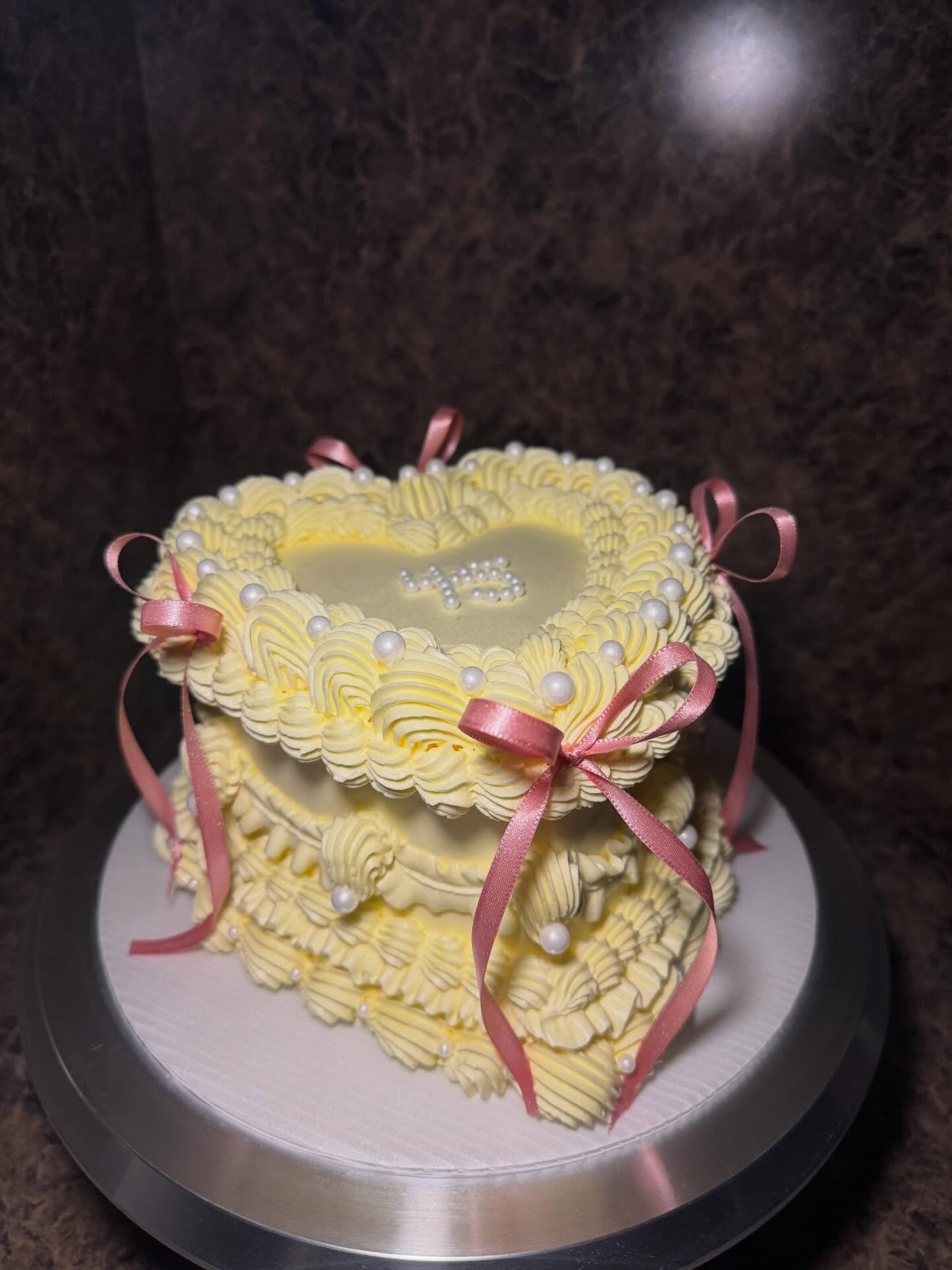 A heart-shaped cake decorated with yellow piped frosting, white pearl-like accents, pink ribbons, and a small white number '13' on top.