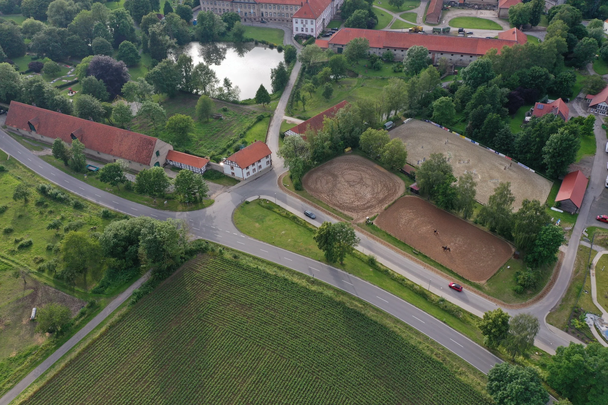 Luftaufnahme eines kleinen Dorfes mit Streuobstwiesen, einem Teich, einem Reitplatz mit Pferden, mehreren Gebäuden, einer Straße und umgeben von grünem Land.