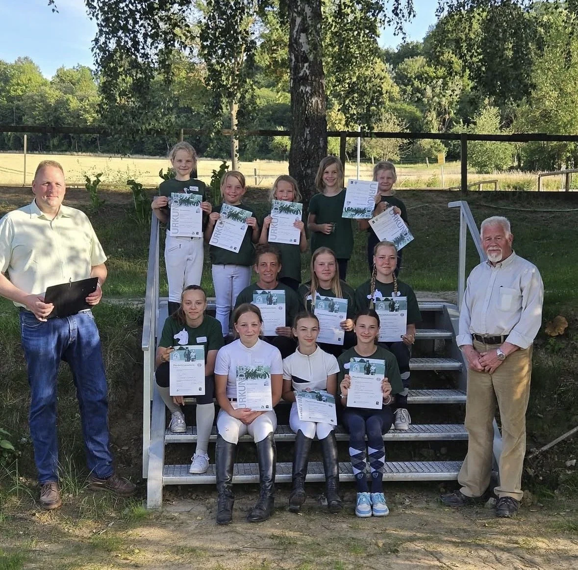 Gruppenfoto von jungen Mädchen, die ihre Urkunden bei einer Veranstaltung im Freien erhalten haben. Sie stehen auf Stufen vor einem Baum mit zwei Erwachsenen, die die Gruppe betreuen.