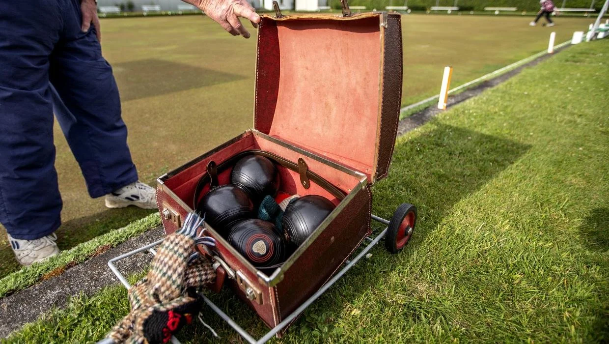 A person holding an open vintage case with four black bowls inside, on the bowling green.