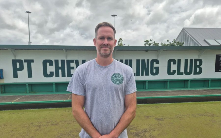Man standing on a bocce court in front of a sign that reads 'PT CHEVY BOWLING CLUB.' The man is wearing a gray t-shirt with a logo on the left chest, and the background features cloudy skies.