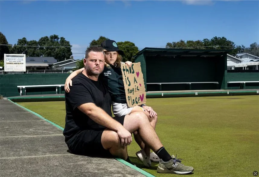 A man and a young girl sitting by the bowling green. The girl is holding a sign that reads 'this is a magic place.'