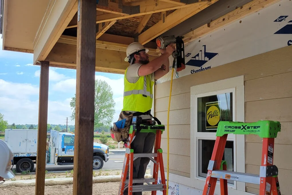 Construction workers in safety vests and helmets framing the interior of a house under construction, with exposed wooden beams and a window.