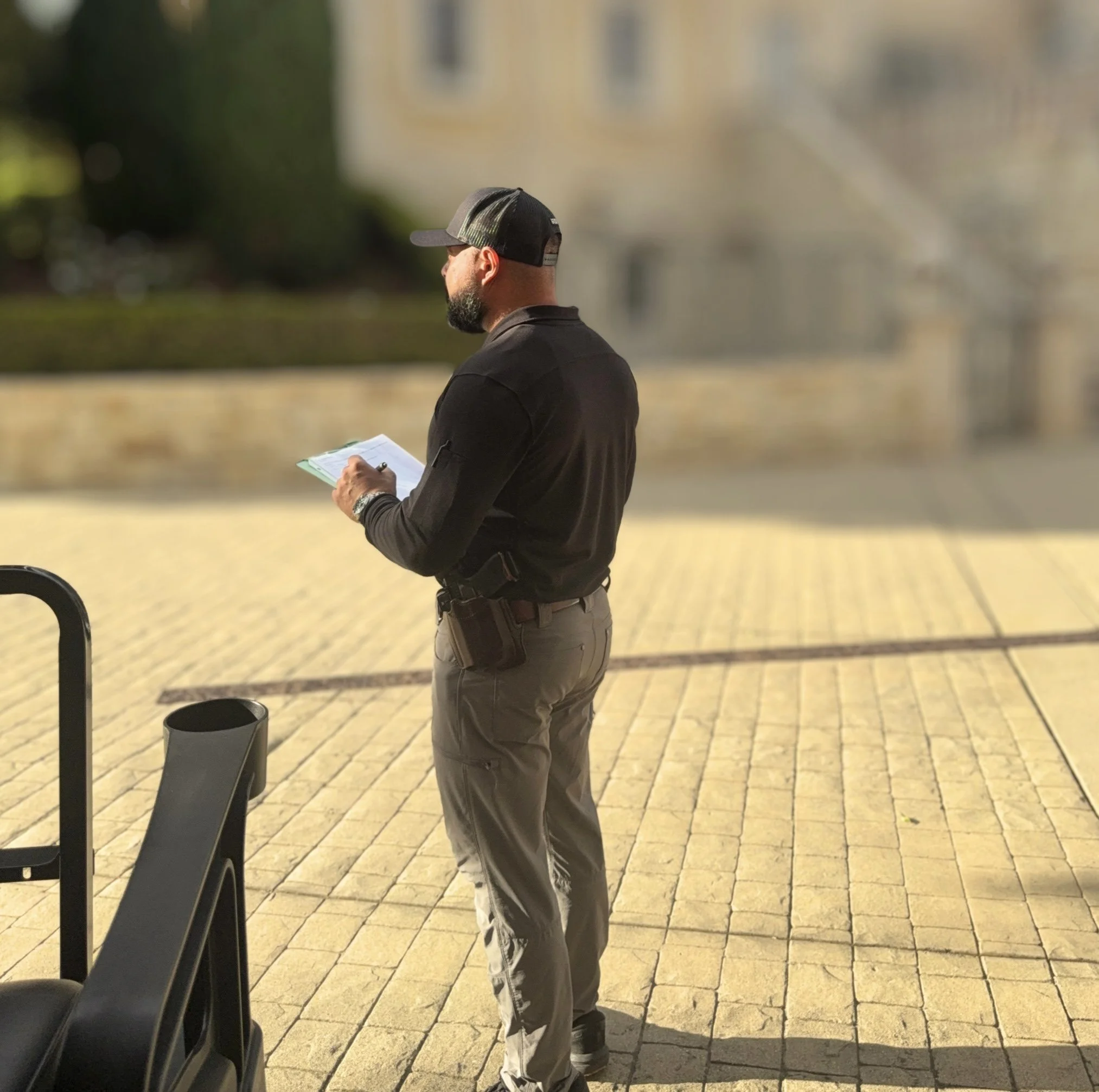 A man wearing a black cap, black jacket, and gray pants reading a paper outdoors near a row of black chairs on a yellow brick pavement.