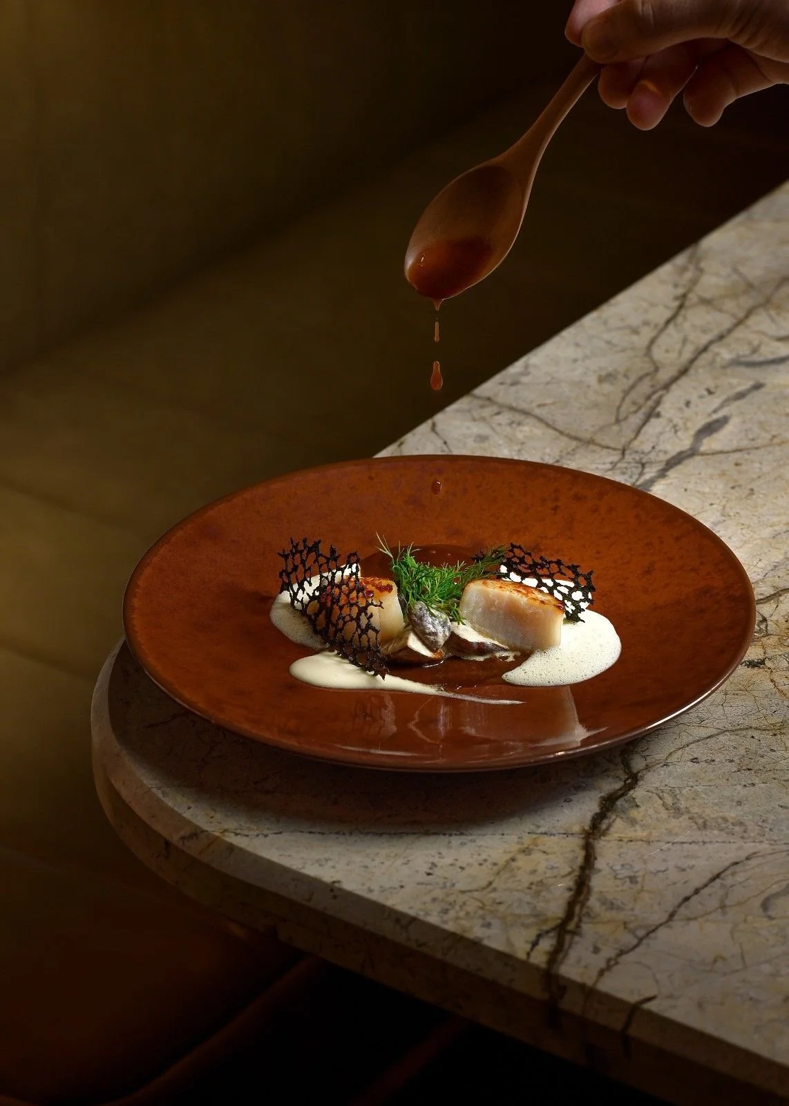 A chef drizzles sauce onto a plated gourmet dish featuring seared scallops, foam, fresh herbs, and decorative black crisps on an orange ceramic plate.