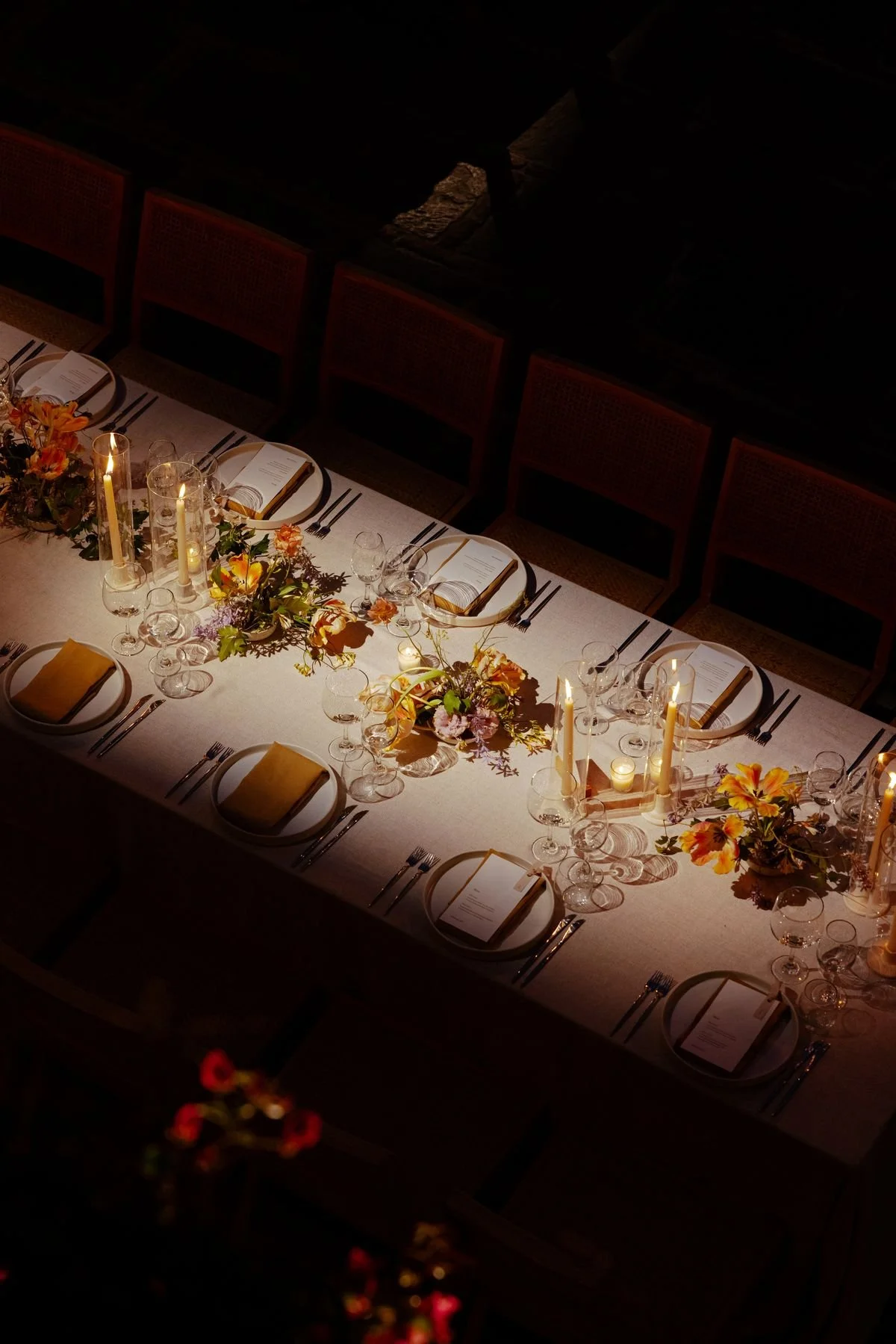 Elegant dining table set for a formal event with floral centerpieces, candles, wine glasses, and place settings, illuminated by soft lighting in a dimly lit room.