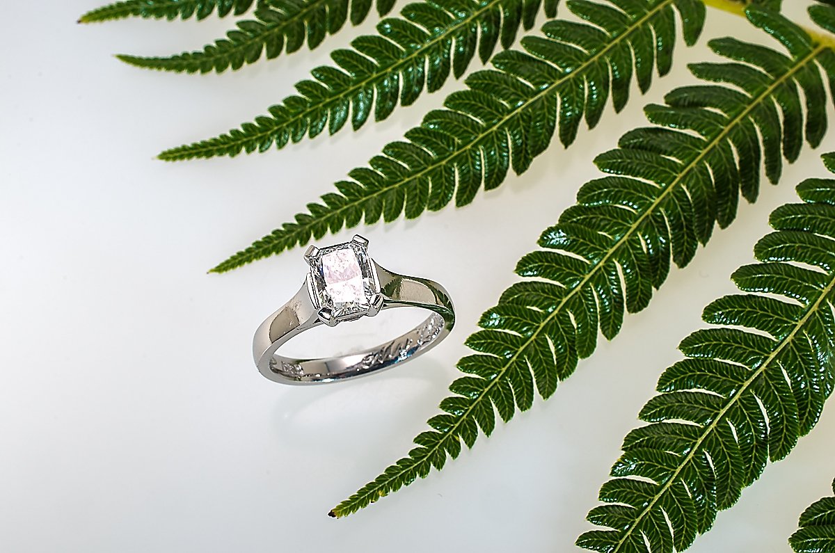 A silver ring with a pink gemstone in a rectangular cut, surrounded by green fern leaves on a white background.