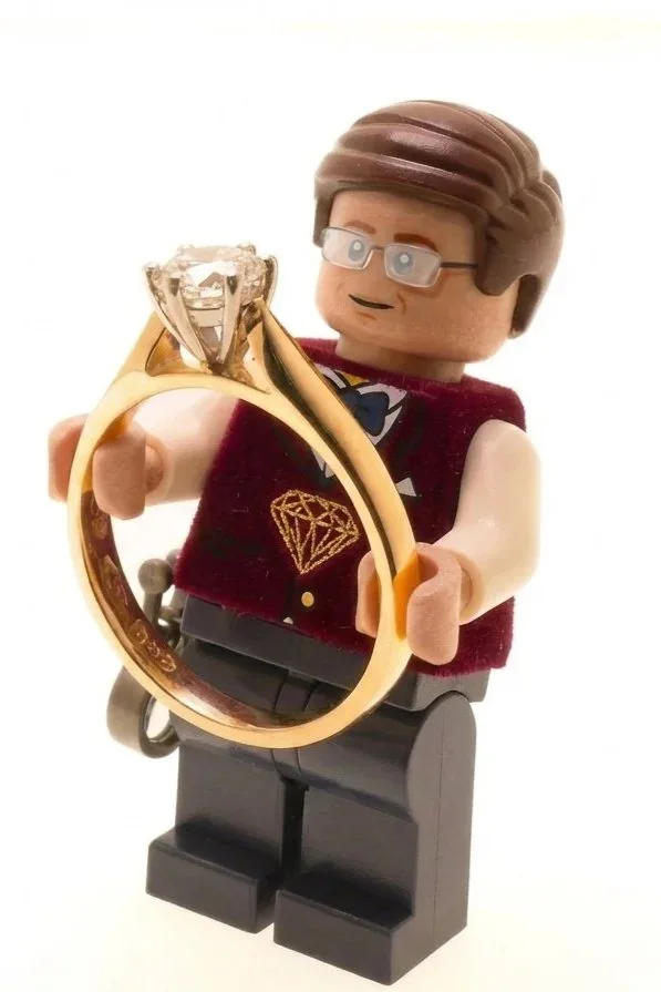 LEGO figurine of a man with glasses, wearing a vest with a diamond design, holding up a large ring with a diamond, against a white background.