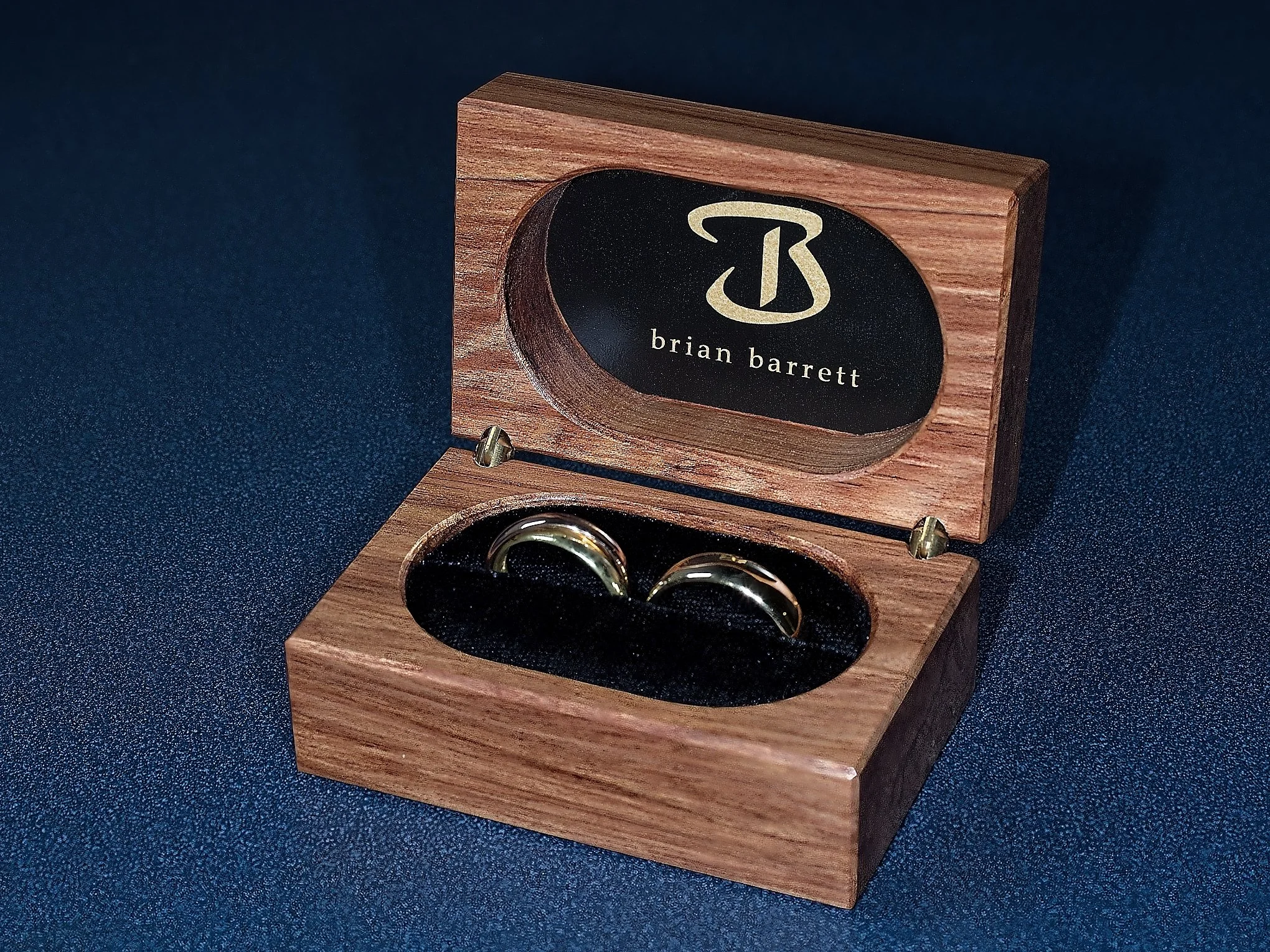 Wooden box with a black interior holding pair of gold wedding rings, lid has a black oval with gold 'B' logo and the name 'brian barrett' in white text.