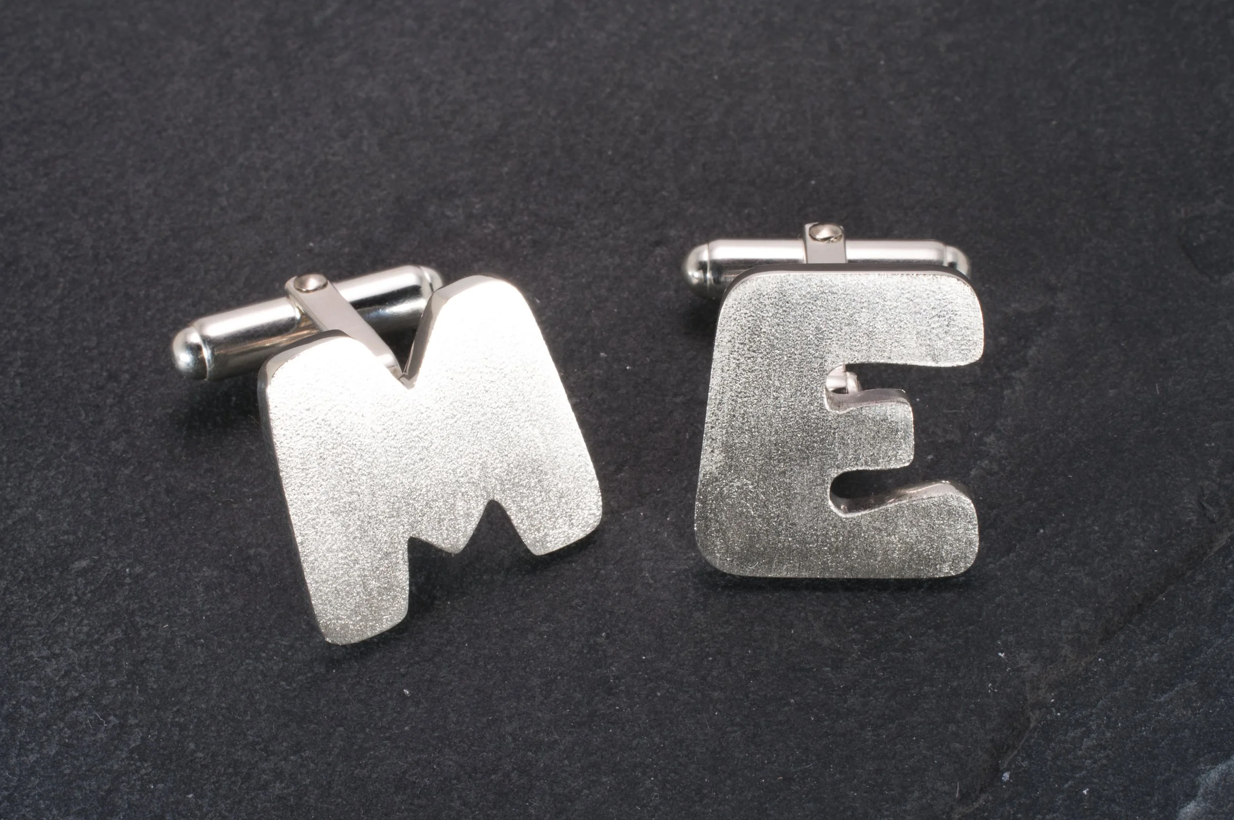 Silver cufflinks shaped like the letters 'M' and 'E' on a black background.