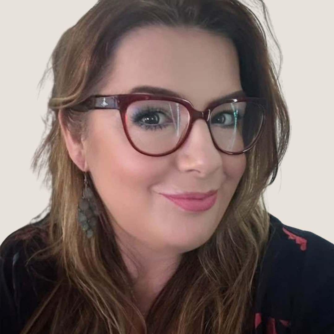 Close-up of a woman with long brown hair, wearing red glasses, makeup, and a black top with red accents, smiling at the camera.
