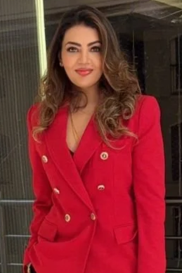 A woman with long wavy hair wearing a red blazer with gold buttons, standing indoors with a window and dark background.