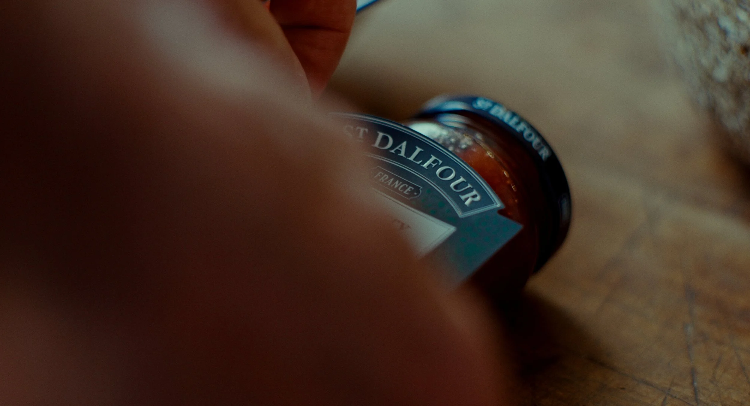 Bouteille de confiture placée sur un sol en bois, avec une main partiellement visible dans le cadre.